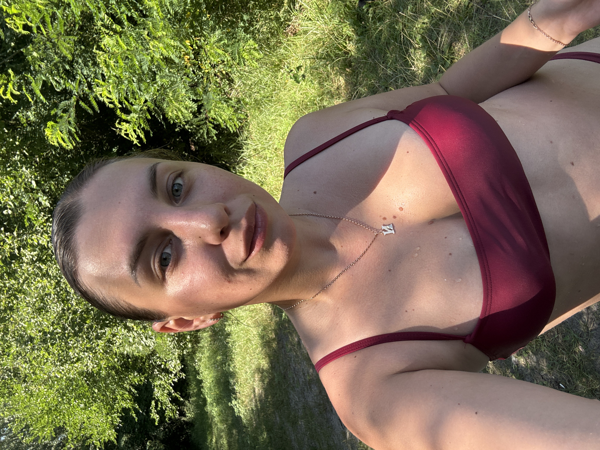 A woman is taking a selfie outdoors. She is wearing a burgundy bikini top and a silver necklace with a pendant. Her hair is pulled back, and she has a light-colored complexion. She is smiling slightly. The background is filled with lush green trees and grass, suggesting a natural outdoor setting, possibly a park or a wooded area. The lighting indicates it is daytime, with sunlight filtering through the foliage. The image appears to have been taken in Chykalivka, Ukraine, given the context.