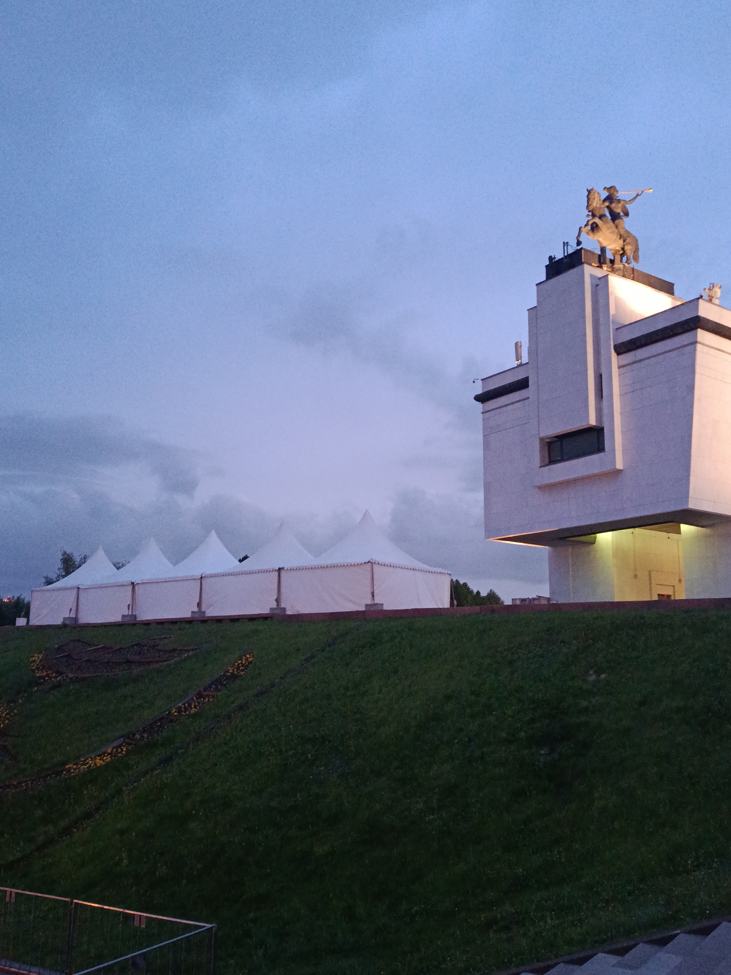 The image shows a view of the Victory Monument in Moscow, Russia, along with several white tents situated behind it. The monument features a white stone building with a statue of St. George slaying the dragon on top. The surrounding area is a green grassy hill, with stairs leading up to the monument from the foreground. The sky is cloudy.