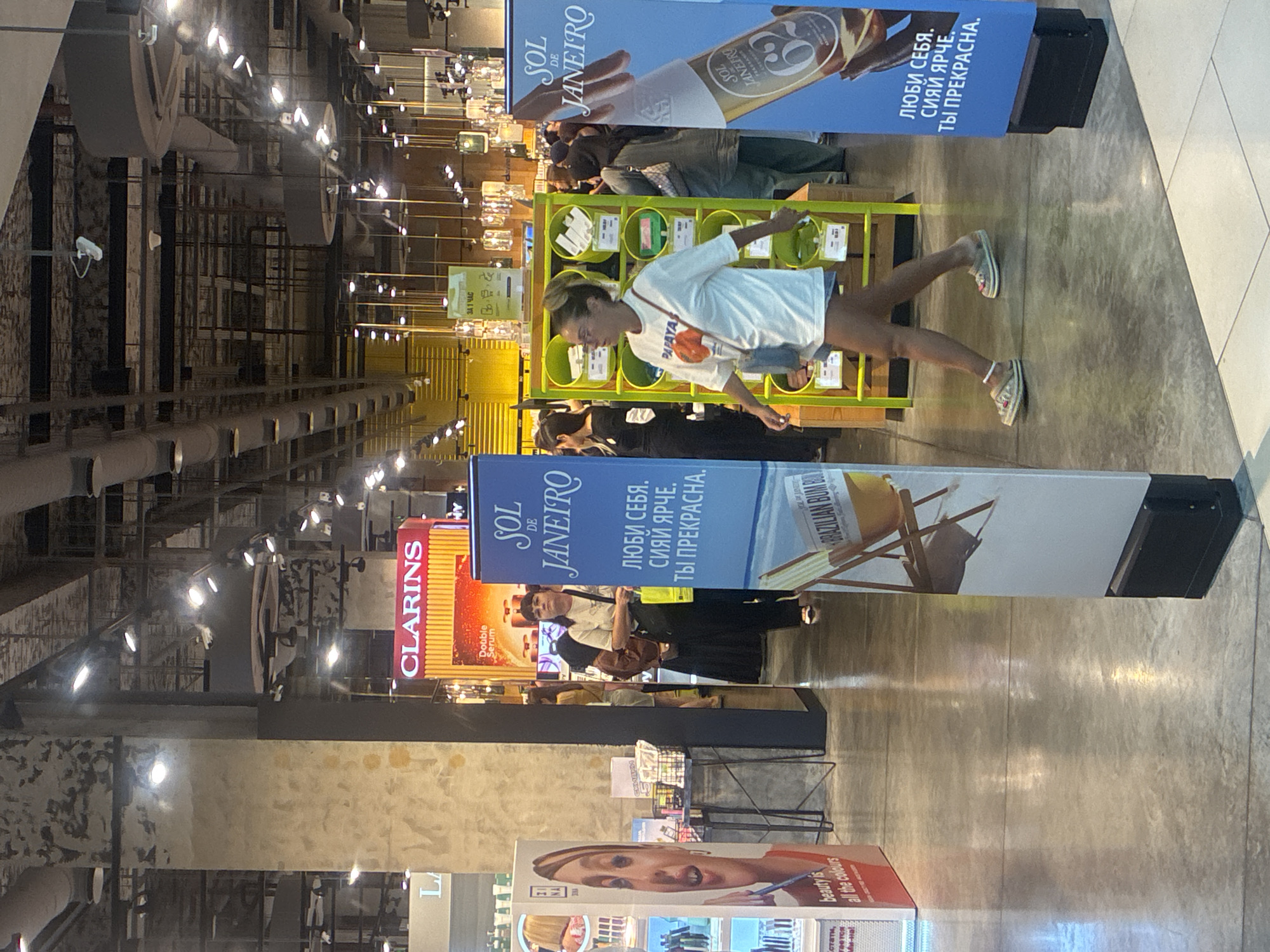 The image depicts an indoor retail environment, likely a department store or a large cosmetics retailer, with a polished concrete floor reflecting overhead lighting. In the foreground, a woman with blonde hair tied back is walking to the right, wearing glasses, a white t-shirt with a graphic print, and denim shorts. She has a small crossbody bag. To her left, a tall blue display sign for "SOL DE JANEIRO" features text in Russian and an image of a product bottle. To the far left, a vertical display promoting "ZI NA BEAUTY" shows a woman's face. In the midground, a woman with dark hair and bangs is standing and looking towards the left, wearing a light-colored top and dark pants, carrying a yellow shopping bag. Behind her, another person in a dark head covering is visible. Further in the background, a display for "CLARINS" with red and orange branding is prominent. The ceiling features industrial elements with exposed pipes and multiple track lights illuminating the space. The overall impression is a well-lit, modern commercial setting.