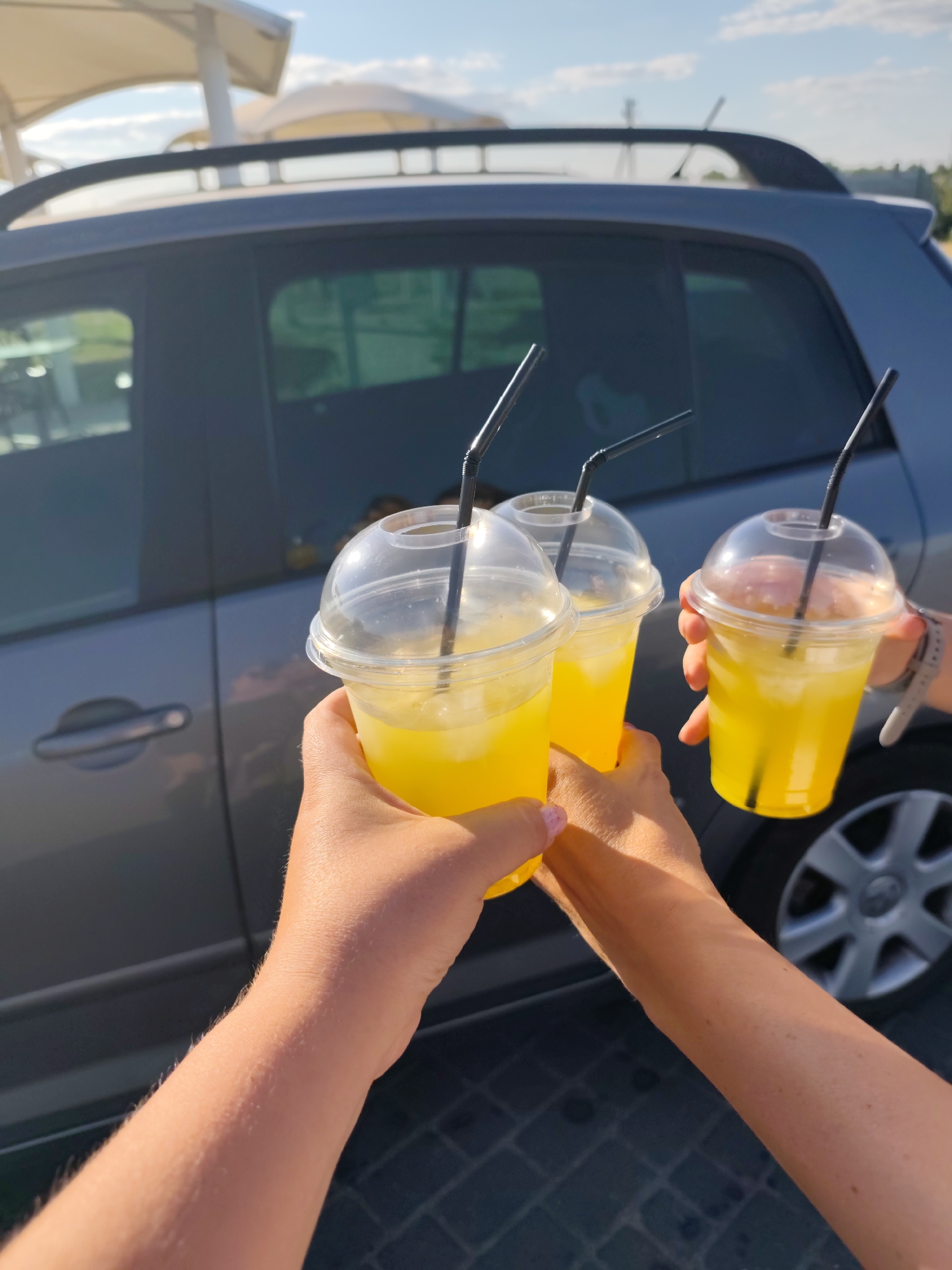 The media file shows three hands holding clear plastic cups filled with yellow liquid and ice, each with a black straw. The cups are likely containing a beverage such as lemonade or iced tea. The background features a gray car with tinted windows, and in the distance, there are structures that appear to be awnings or shelters, suggesting an outdoor setting. The lighting indicates it is daytime, with bright sunlight casting shadows. The hands are varied in appearance, with one hand showing painted fingernails, and another with visible arm hair, suggesting multiple people are present. The overall mood conveyed is casual and celebratory, possibly a toast or a moment of enjoyment. There is no visible text in the image. The provided location context is Nechaiane, Ukraine.