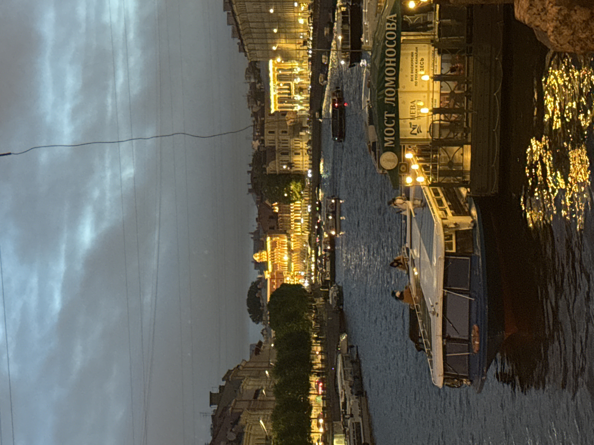 The image depicts a canal in Saint Petersburg, Russia, at night. Buildings line the canal, illuminated with warm light. Boats are docked along the canal, including one labeled "Most Lomonosova" (Lomonosov Bridge). There are a couple of people visible on one boat. The sky is overcast and the water reflects the city lights.