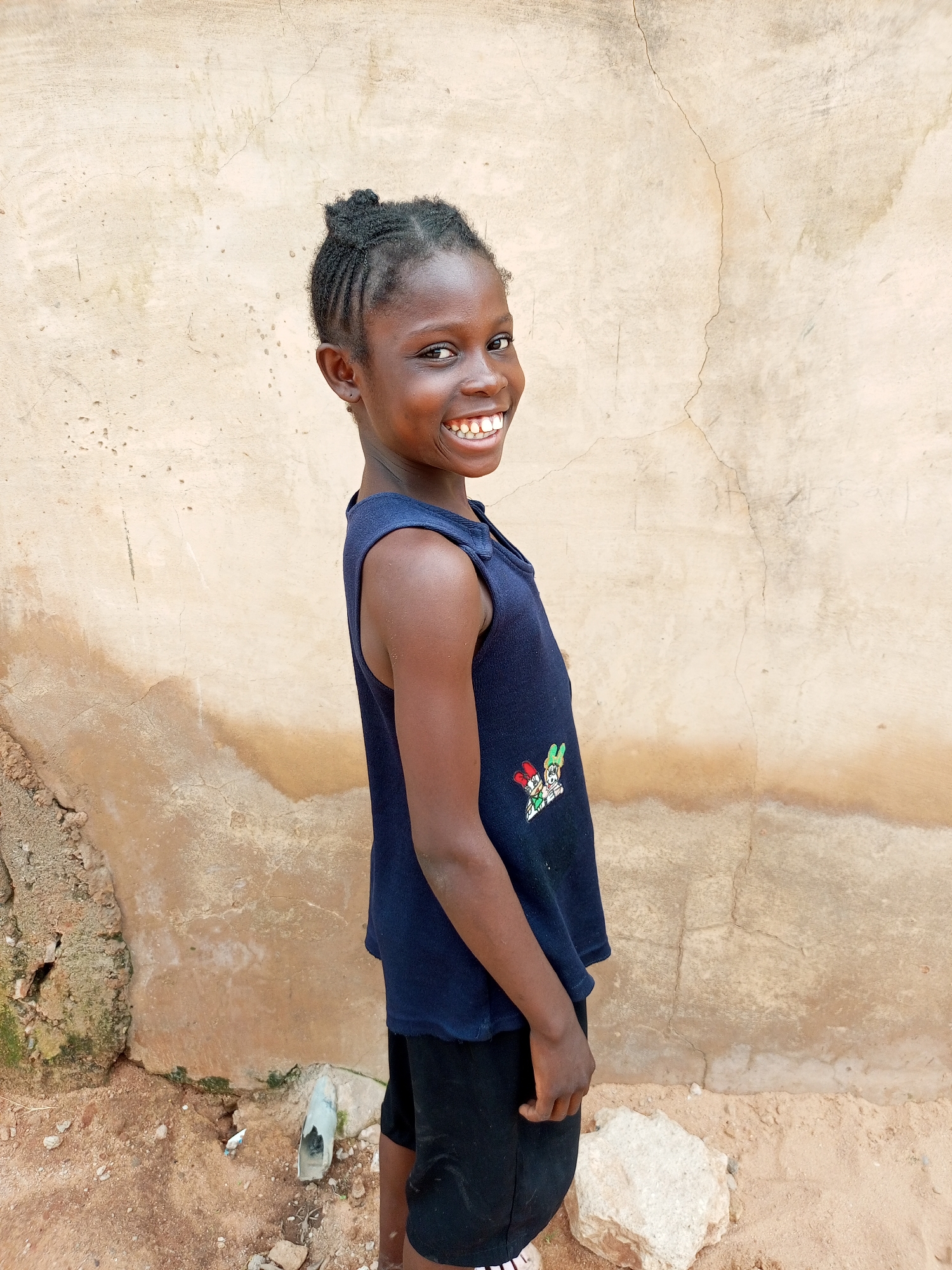 A young girl with braided hair pulled into a bun stands facing a textured, light-brown wall. She is wearing a navy blue sleeveless shirt with a cartoon design on the front and black shorts. She is smiling widely and looking over her left shoulder towards the camera. The ground in front of the wall is sandy and contains some rocks and debris, including a white plastic object. The wall appears weathered with cracks and discoloration. The scene appears to be outdoors in what could be a rural or semi-urban area, possibly in Tempure, Nigeria.