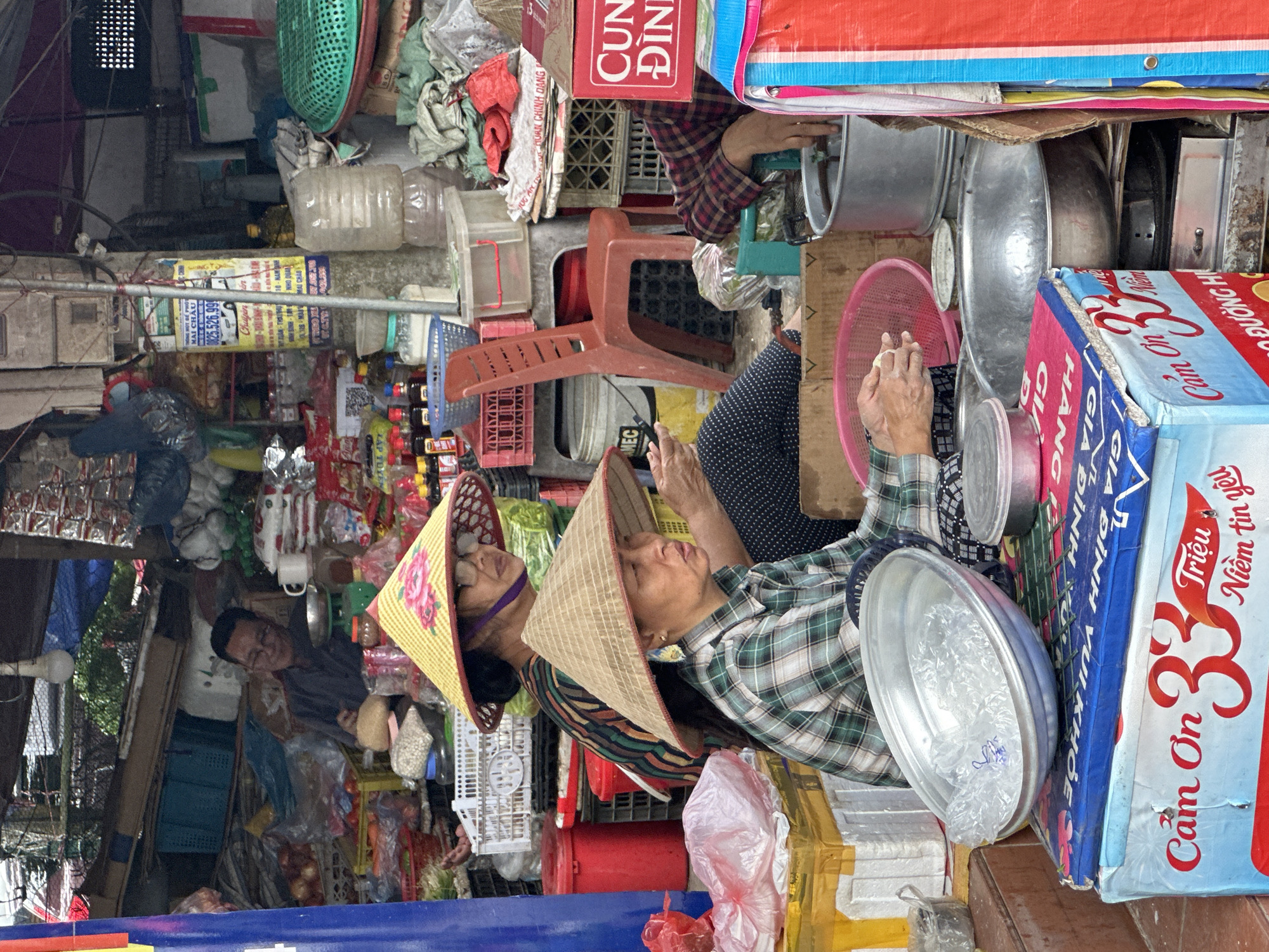 A street market scene is depicted in Mai Chau, Vietnam. Two women are central to the image, both wearing traditional conical hats. The woman in the foreground, with a light brown hat, wears a green and white plaid shirt and is looking down, her hands engaged with something unseen. Behind her, a second woman with a yellow hat adorned with pink flowers wears a dark, striped shirt and glasses, looking off to the right.
The market environment is cluttered with various goods. Crates and containers filled with produce, plastic bags, and bottles are visible. A red plastic chair is positioned centrally, and a red cylindrical container is to the left. In the background, a man in a dark shirt and glasses is partially visible, appearing to be working at a stall. The overall impression is one of a busy, outdoor marketplace with an abundance of merchandise.