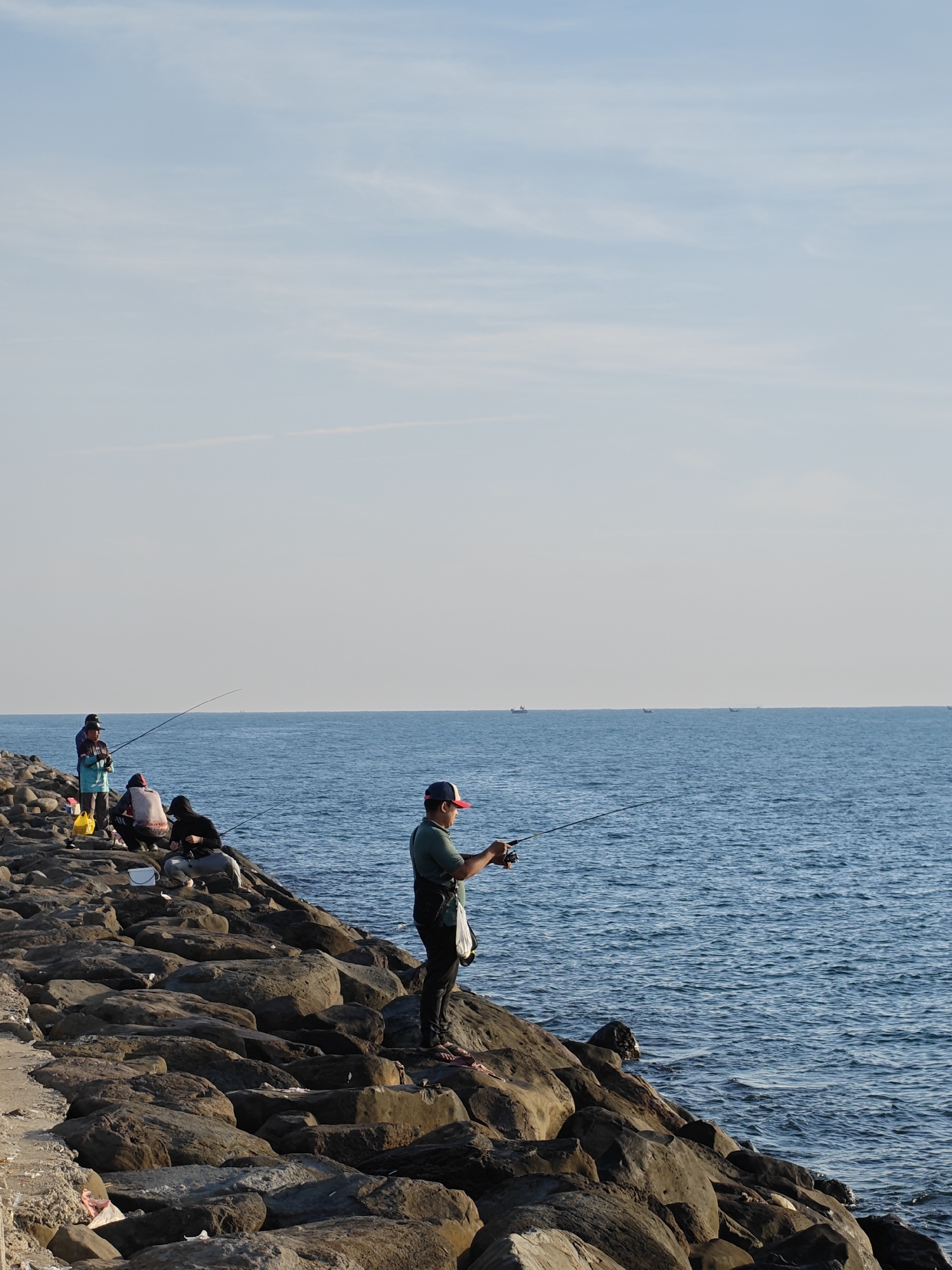 The image shows a group of people fishing from a rocky breakwater along the coast in Lhokseumawe, Indonesia. Several individuals are visible, some standing and casting their lines into the sea, while others are seated on the rocks. The sea stretches to the horizon under a clear sky. The breakwater is composed of large, irregularly shaped rocks. In the distance, the silhouettes of small boats can be seen on the water. The scene is bathed in sunlight, suggesting it is daytime.