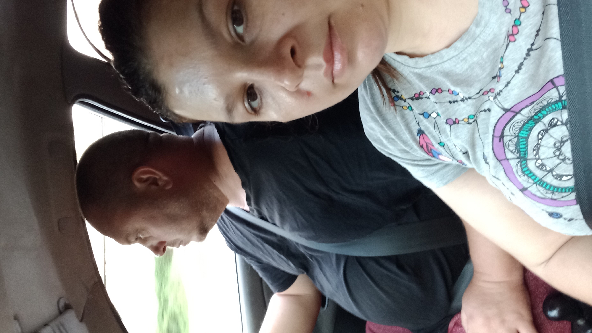 This image shows two people in a car, likely in Kropyvnytskyi, Ukraine. The driver, a bald man wearing a black t-shirt and a seatbelt, is looking to his left. Beside him, a woman with dark hair pulled back is looking at the camera. She is wearing a gray t-shirt with a colorful, bohemian-style print featuring feathers and geometric patterns. The car's interior is visible, including the dashboard and the roof lining. The scene appears to be captured during daytime, with natural light filtering into the car. The background outside the car windows shows a blurred green landscape, suggesting the vehicle is in motion, possibly on a road outside the city center or on the outskirts of Kropyvnytskyi.