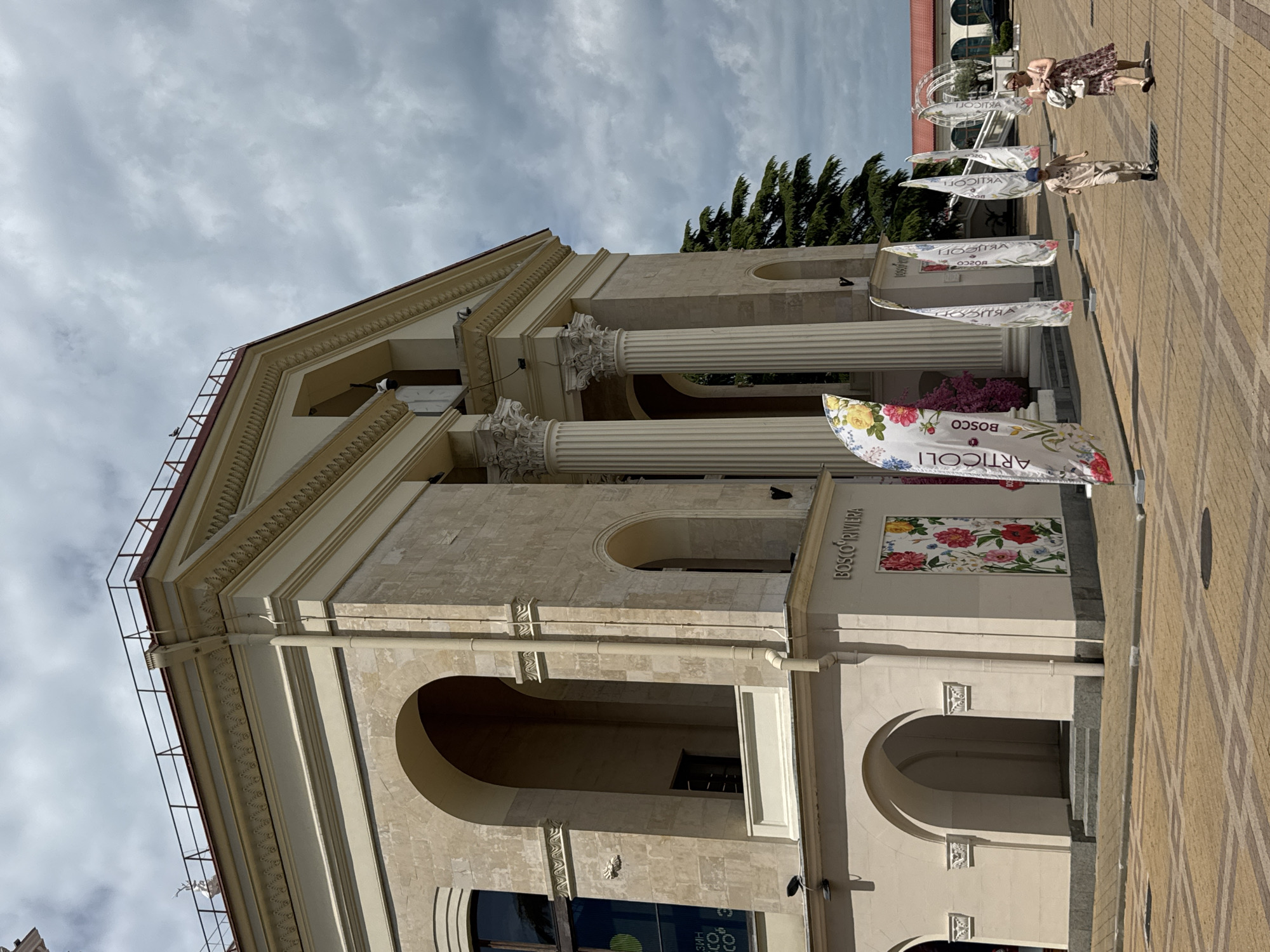 The image shows a building in Sochi, Russia, recognizable by the "BOSCO RIVIERA" sign and distinctive architecture. The building is light-colored and features classical elements such as columns and arches. There are several flags displaying advertisements, including "ARTICOLI BOSCO," positioned along the building's facade. Two people are walking on the paved area in front of the building. The sky is overcast, and there are trees visible in the background.
