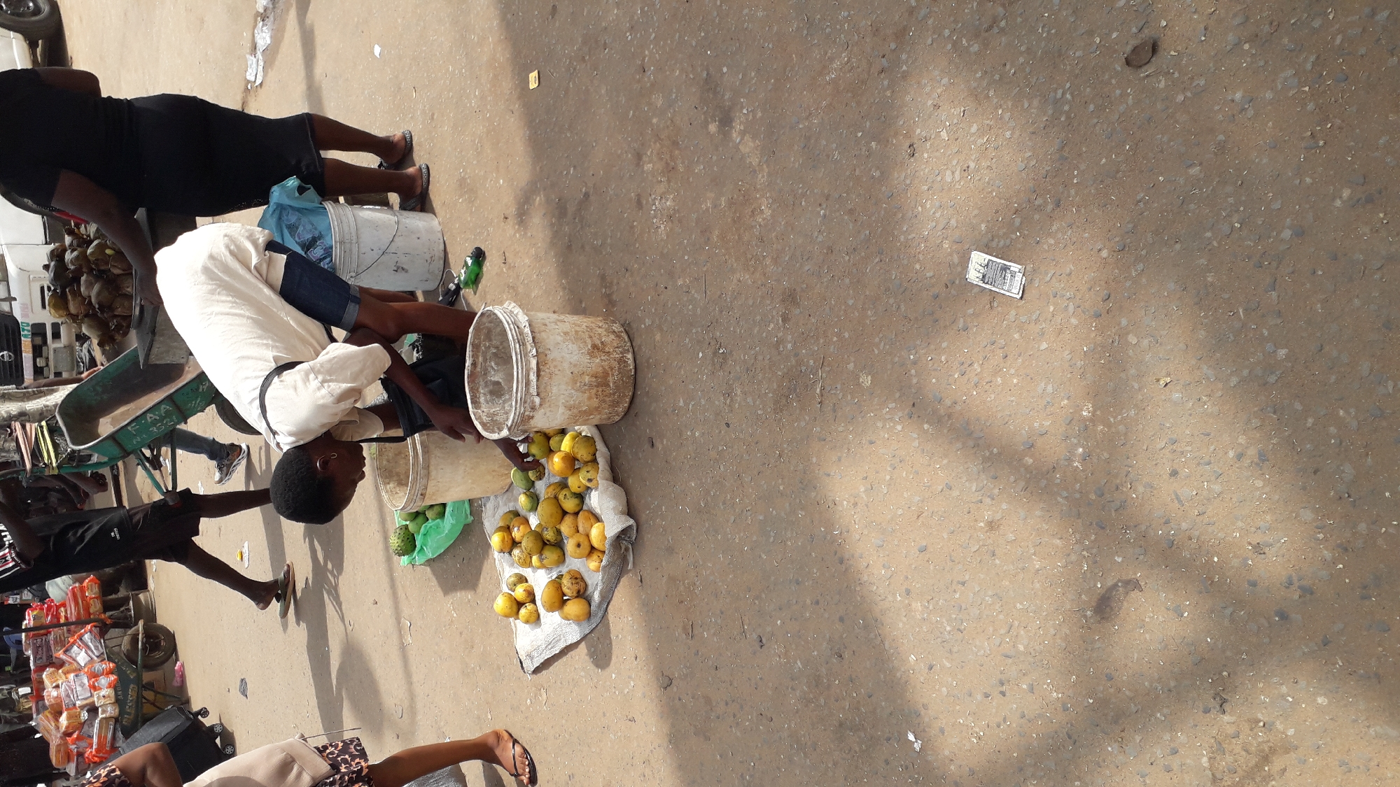 The image captures a street scene in Onitsha, Nigeria, likely a market area. In the foreground, a young woman with short dark hair, wearing a light-colored t-shirt and denim shorts, is bent over a display of fruits on a white cloth. Several yellow fruits, possibly mangoes, are spread out, along with a few green fruits, possibly soursop. Two weathered buckets, one light-colored and the other a dirty white, are placed near the fruits. Another person, a woman wearing a black dress, stands behind the fruit display, partially obscured. In the background, other people are visible, including a man in black shorts and a t-shirt. A green wheelbarrow filled with items is also present, along with a white van and piles of coconuts. The ground is paved with a rough, sandy-colored surface, and shadows from overhead structures indicate an outdoor setting. A small piece of paper, possibly a receipt or ticket, lies on the ground in the lower right portion of the image.