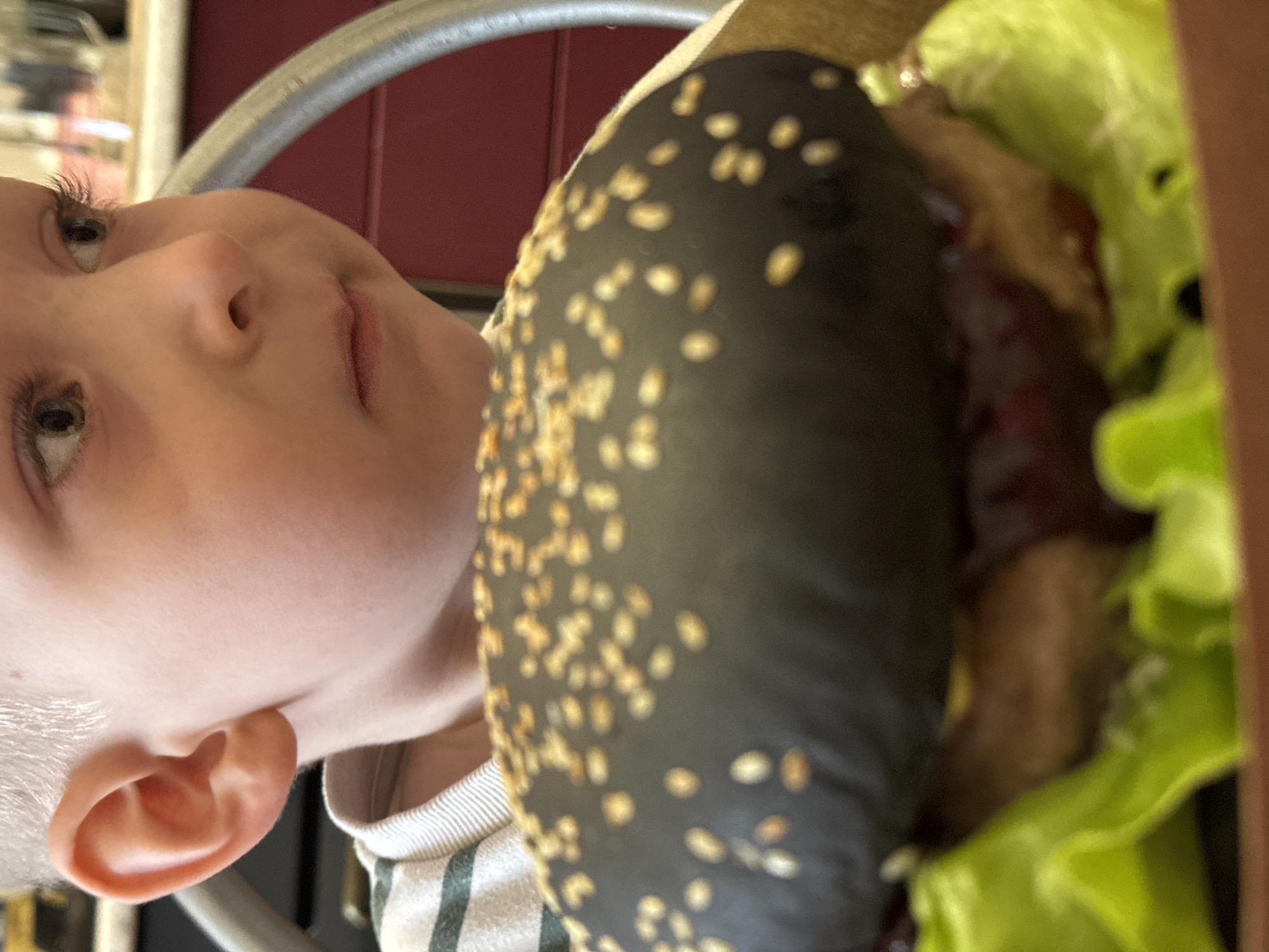 The image is a rotated, close-up shot primarily featuring a young child observing a hamburger, likely in an indoor setting.
On the left side of the frame, a fair-skinned child, appearing to be a baby or toddler with very light hair, is positioned with their head tilted. Their large eyes are looking upwards and slightly to the right, conveying a sense of curiosity or attentiveness. The child's nose and part of their mouth are visible, and a portion of their left ear is also clearly seen. They are wearing a garment with horizontal stripes.
The prominent object in the center-right and foreground is a hamburger. It features a distinctive dark, possibly black or charcoal, bun generously sprinkled with sesame seeds. Beneath the bun, bright green lettuce is visible, along with glimpses of dark reddish-brown contents, suggesting a meat patty and perhaps a sauce.
In the background, a reddish-brown surface, possibly a wall or part of a chair, is visible behind the child's head and the hamburger. A curved metallic grey bar, which could be part of a high chair, a table, or other furniture, runs across the upper portion of the frame. The lighting is bright and appears to be indoor, suggesting the image was taken during daytime. There is no visible text in the image. The location is Perm, Russia.