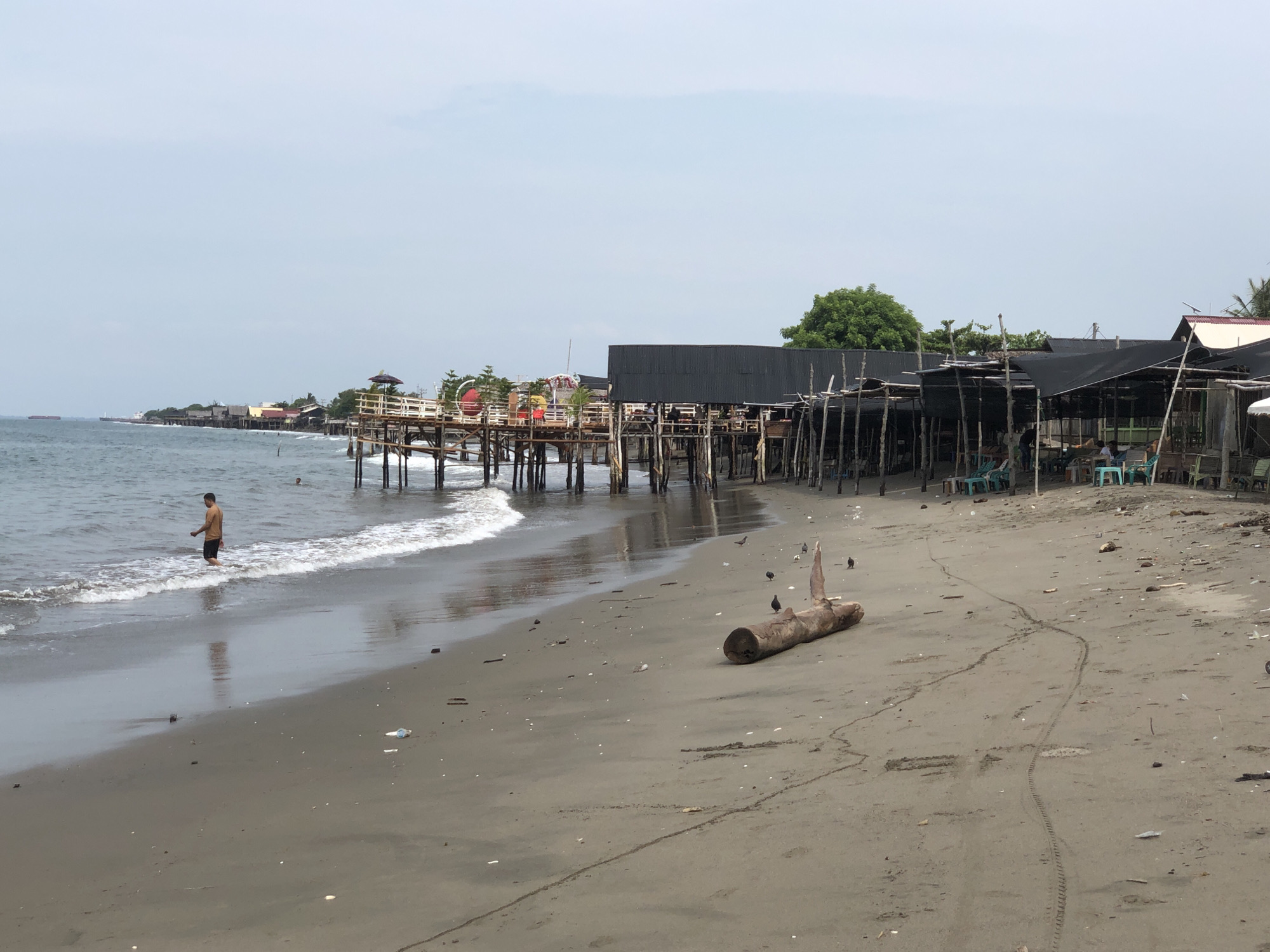 The image depicts a beachfront scene at a location identified as Ulee Blang, Lhokseumawe, Indonesia. The foreground is a sandy beach with some debris scattered, including a large piece of driftwood and several birds. In the mid-ground, there is a wooden pier with several structures built on it, covered with black tarps. Some people are seated at tables under these structures, suggesting a dining or seating area. The ocean is to the left, with gentle waves lapping at the shore. A single person, wearing an orange shirt and black shorts, is standing in the shallow water. The sky is overcast and a pale blue. In the background, more structures can be seen along the coastline, and two ships are visible on the horizon. The overall impression is that of a casual, potentially tourist-oriented beach area.