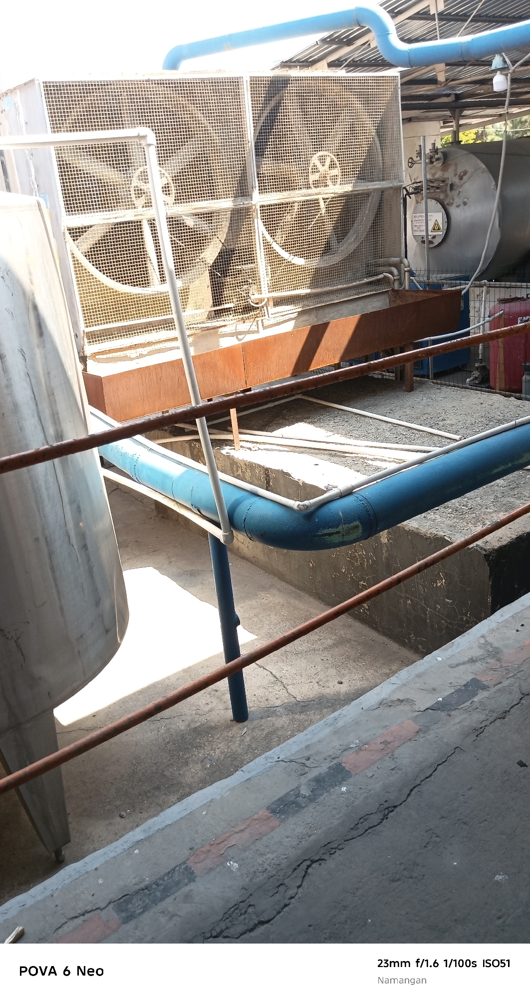 This image, taken in Namangan City, Uzbekistan, shows a large industrial fan unit with its protective mesh coverings. Two large fan blades are visible behind the mesh on the left. In the background, a cylindrical metal tank with a warning label is present. A blue pipe runs horizontally across the scene, supported by white pipes and a blue vertical pole. The surrounding environment consists of concrete ground and industrial structures, with some visible rust and wear on metal components. The lower portion of the image shows a cracked concrete surface with painted lines.