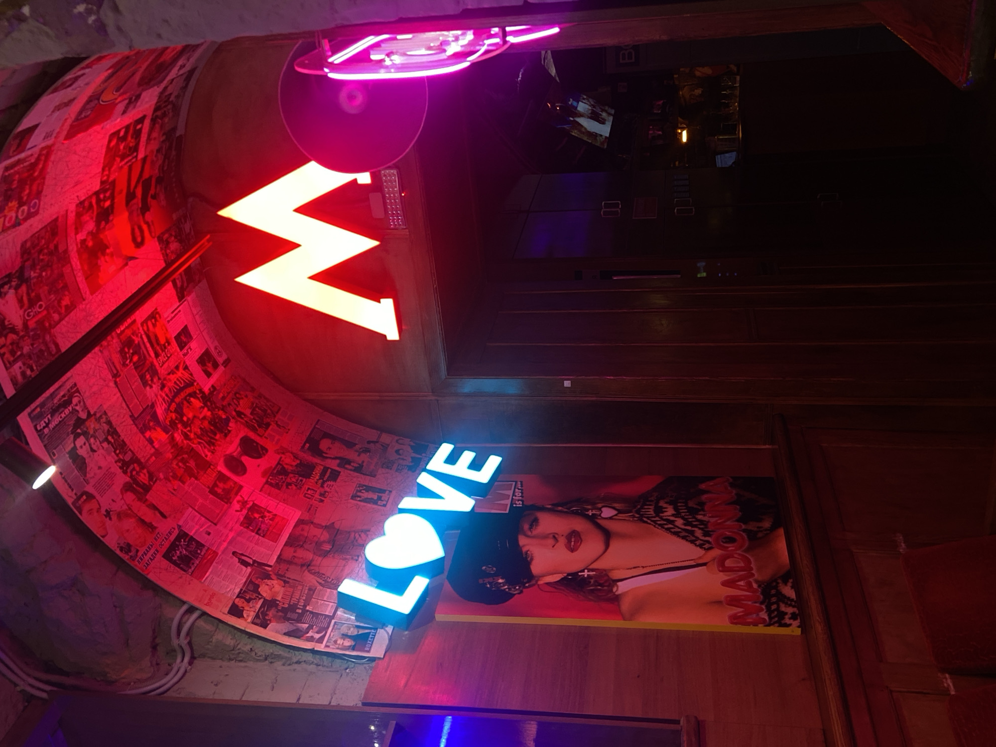This image shows an interior space decorated with vibrant neon lighting and pop culture references. In the foreground, the word "LOVE" is spelled out in bright blue neon letters, with a heart symbol replacing the "O." Behind the neon sign is a poster of the singer Madonna. A large, red neon arrow is also visible, pointing towards a ceiling covered in collages of newspaper clippings and other images. The overall effect is bold and visually stimulating, suggesting a trendy or themed establishment. The image is taken in Saratov, Russia.