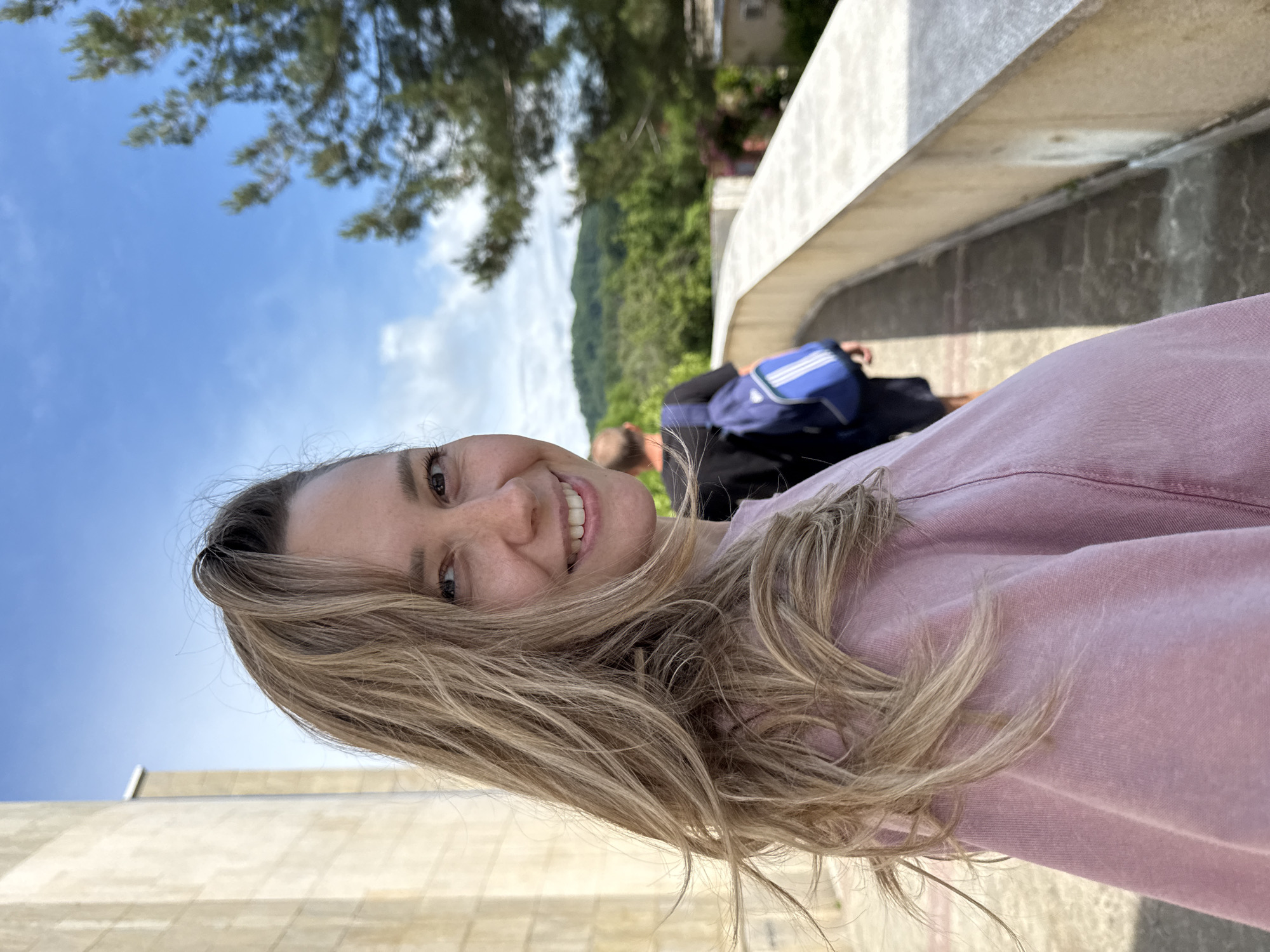 In the city of Nebug, Russia, a woman with blonde hair is captured in a close-up selfie, looking over her shoulder and smiling at the camera. She is wearing a pink shirt and her hair is blowing in the wind. Behind her, a man with a blue backpack walks away from the camera down a path bordered by a stone wall. The background features lush green trees and hills under a bright blue sky.