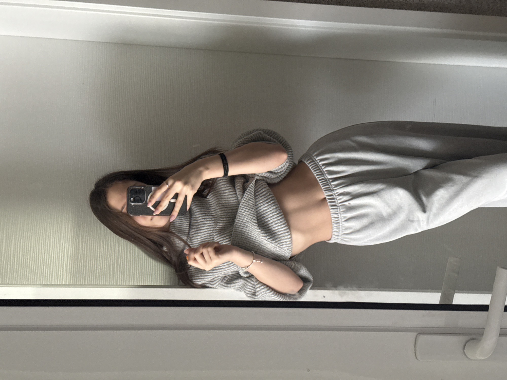 A young woman is taking a mirror selfie, standing indoors. She is wearing grey sweatpants and a grey striped sweater that exposes her midriff. She holds a black iPhone in her right hand, obscuring her face. She has long brown hair and is wearing a black bracelet on her right wrist and a silver bracelet on her left wrist. The background shows a white wall and part of a window frame with a white handle. The location cannot be verified as Sofia, Bulgaria.