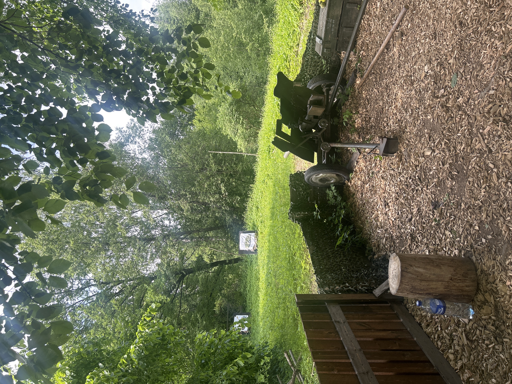 The image depicts an outdoor scene with elements that suggest a historical reenactment or training area. In the foreground, there is a wooden fence and a large tree stump with a bottle of water next to it. A military-style cannon is positioned behind a camouflage net, with ammunition crates visible to the right. The ground is covered in wood chips. In the background, a grassy field is dotted with trees, and two targets, possibly for archery or shooting practice, are visible. The overall environment is lush and green, indicating a natural setting. The presence of the cannon and targets suggests a location related to military history or activities. It is plausible this scene is located in Shlisselburg, Russia, a city known for its historical significance, particularly its fortress.