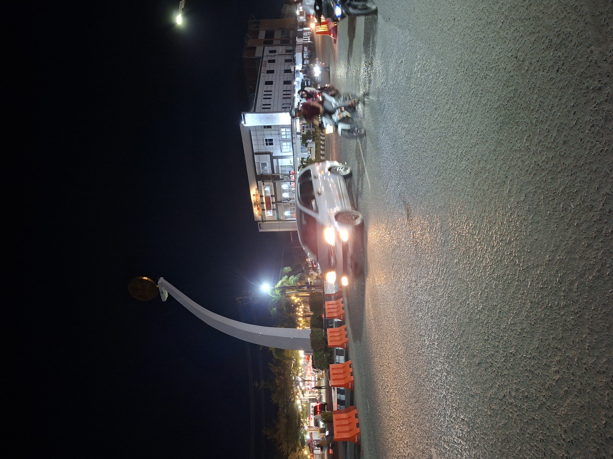 The image captures a night scene in Lhokseumawe, Indonesia. A road occupies the foreground with a visible surface texture. Several vehicles, including a white car and motorbikes, are moving along the road. The car has its headlights on, creating light streaks. Adjacent to the road, orange barriers are placed. In the background, buildings with lit windows are visible. Additionally, there's a large, white, arched structure that appears to be a monument with a circular object on top. The sky is dark, suggesting it's nighttime. The scene is illuminated by streetlights and the lights from buildings and vehicles.