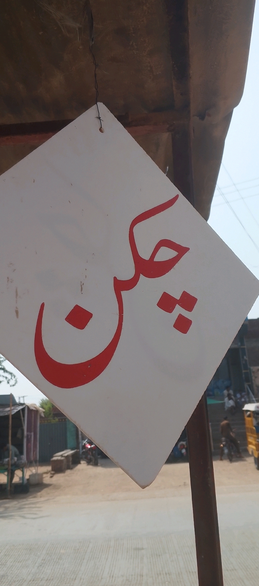 A white sign with red Urdu script hangs diagonally from a rustic, brown corrugated metal overhang. The script, appearing to say "چکن" (chicken), is bold and stylized. Below and behind the sign, a dusty street is visible, lined with small shops and stalls. Several motorcycles are parked or in motion, and a few people are seen in the background, some appearing to be customers or vendors. The overall setting suggests a bustling marketplace or roadside food stall in a town like Lilla, Pakistan.