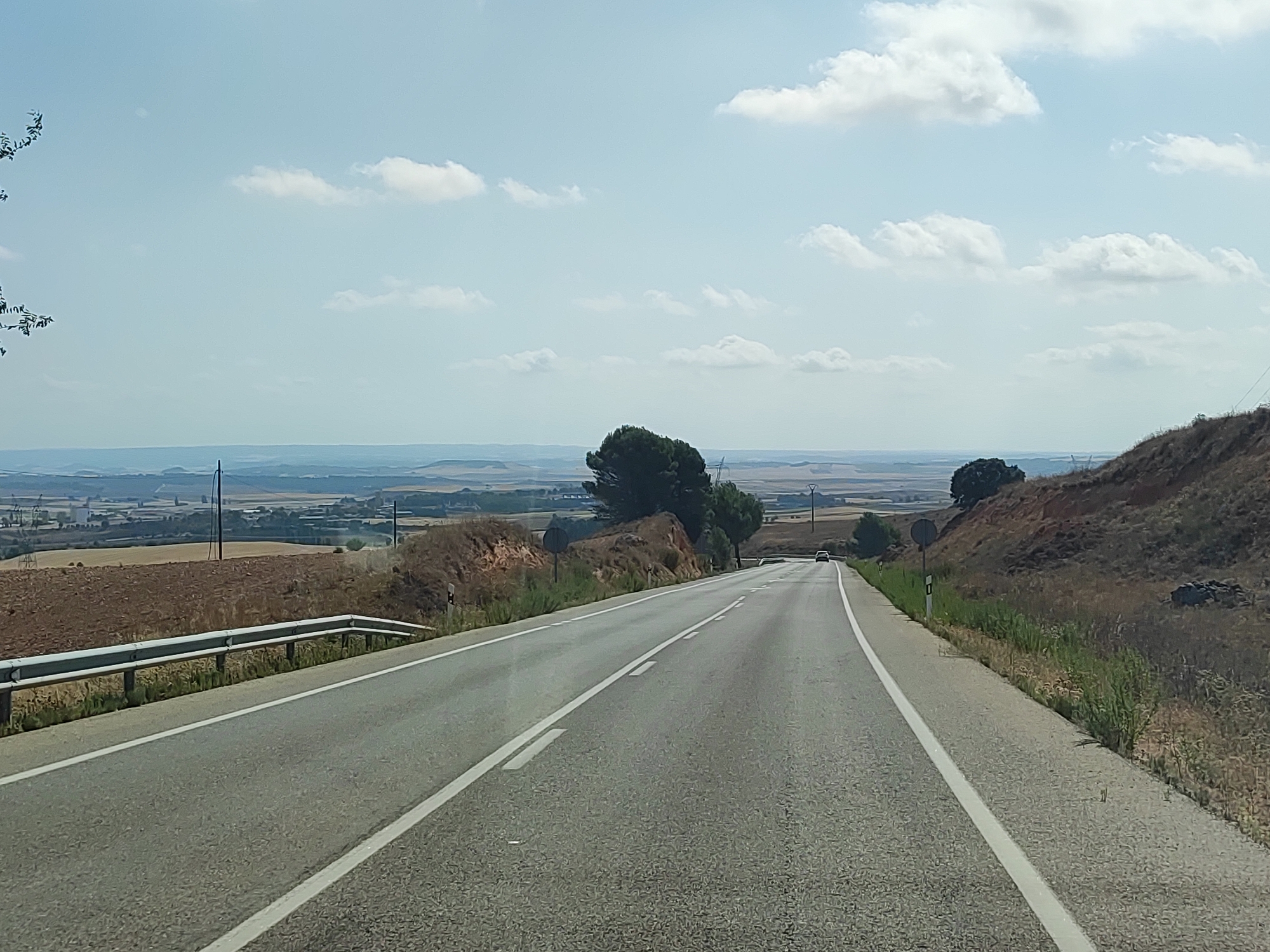 A paved two-lane road curves gently to the right, leading into the distance under a hazy, pale blue sky dotted with white clouds. The road is bordered by a silver metal guardrail on the left and dry, grassy hillsides on the right. In the far distance, a flat landscape of agricultural fields, scattered trees, and some buildings can be seen. The image appears to have been taken from a moving vehicle, likely in a rural area near Castillo de Garcimuñoz, Spain, given the arid vegetation and landscape. There are no people visible in the image.