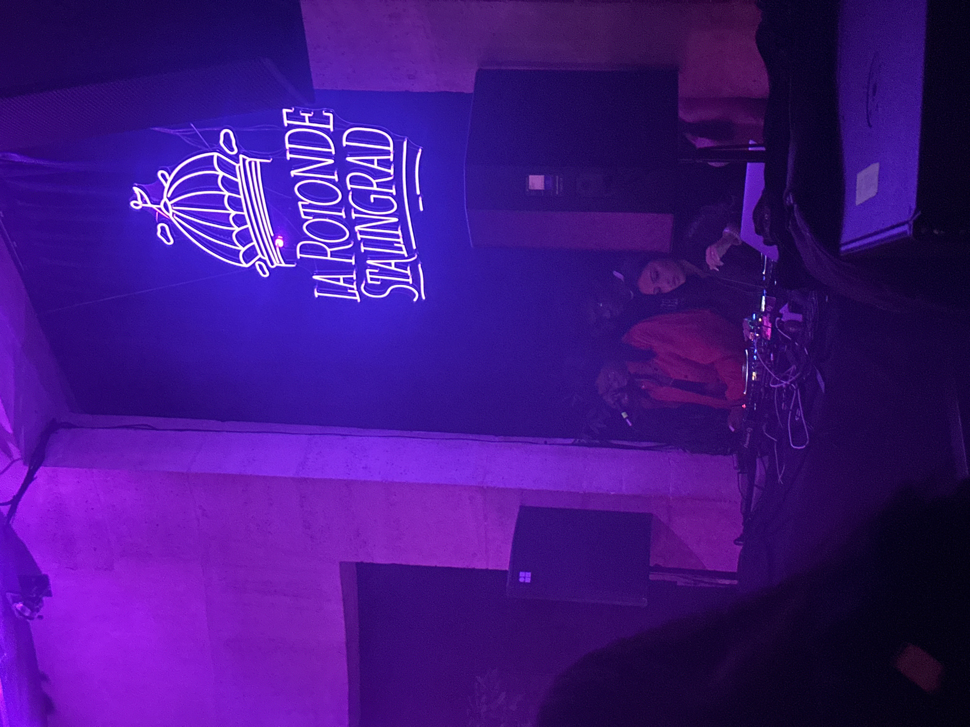 The image depicts the interior of the La Rotonde Stalingrad, a venue located in Paris, France. A neon sign bearing the venue's name and an image of a rotunda is prominently displayed. Two individuals are visible on stage; one appears to be a DJ, positioned behind equipment, and the other seems to be a performer with a microphone. Speakers and other stage equipment are also visible. The lighting is predominantly purple, contributing to the atmosphere of the scene.