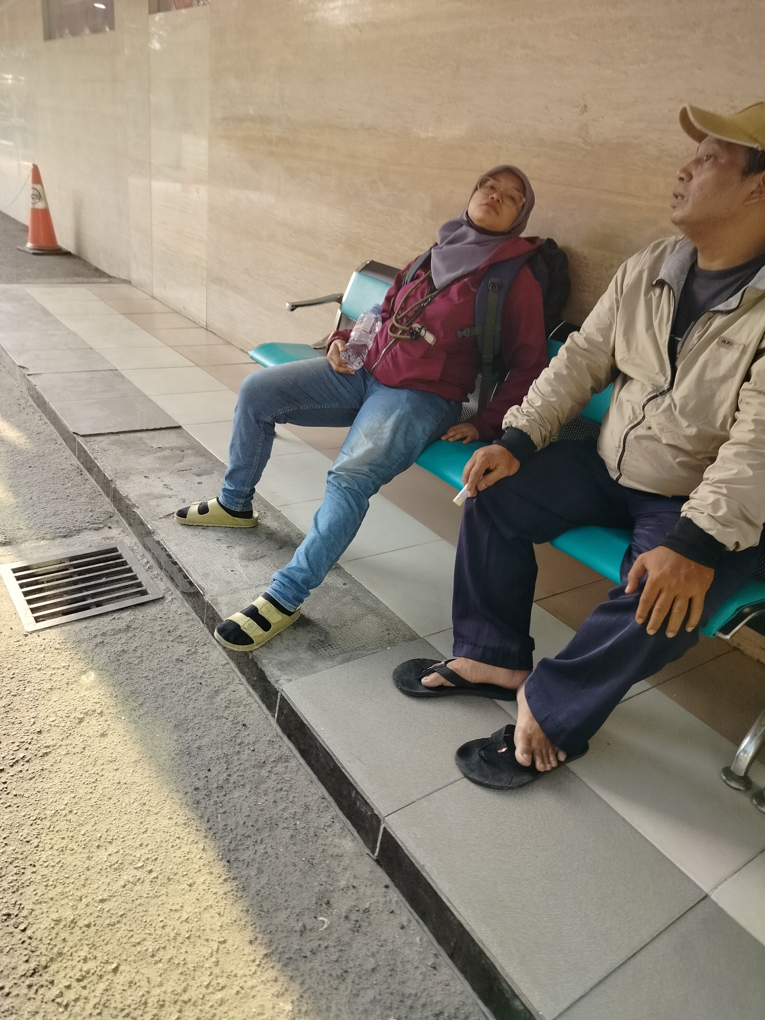 In East Jakarta, Indonesia, two individuals are seated on a teal bench, awaiting something. The person on the left, wearing a purple hijab and a maroon jacket over blue jeans and yellow sandals, holds a bottle of water. To their right, a man in a beige jacket, dark pants, and a tan baseball cap looks upward. His hand rests on his knee, and he appears to be holding a cigarette. The scene takes place outdoors, with a textured wall and a drainage grate in the foreground.