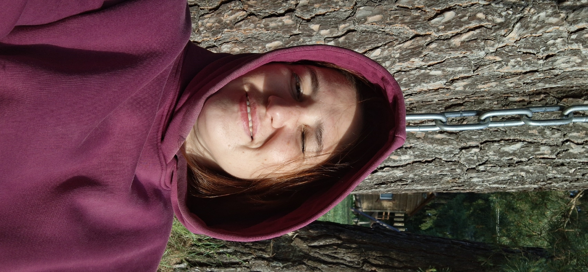 A woman wearing a maroon hooded sweatshirt is lying on what appears to be a tree trunk. A metal chain is visible against the tree trunk behind her head. In the background, there is a glimpse of a wooden building and green foliage. The scene appears to be outdoors, possibly in a forested area. The location is said to be Прикамский, Russia.