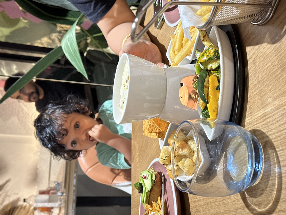 The image depicts an indoor dining setting within a restaurant or cafe in Kalyan-Dombivli, India. A light-colored wooden table occupies the foreground, supporting various food items and tableware. A clear glass with a blue base sits on the lower-left. Centrally, a white ceramic fondue set is present, with a lit candle warming a bowl of light-colored, creamy substance topped with small, dark particulates. Surrounding the fondue are multiple white dishes containing distinct food items: cubed, seasoned bread, triangular corn chips, french fries (some in a small metal basket, others in a dish), and a serving of cooked vegetables including broccoli, sliced yellow and green squash, and baby corn. To the left, on a pink plate, a piece of toast is topped with a green spread, a small dark sphere, and a red spherical element.
A young child with dark, curly hair, wearing a light green t-shirt, is seated in a light brown booster seat behind the table, looking towards the viewer with fingers inserted into their mouth. To the right, an adult's hand, adorned with a gold bracelet, is holding the upper portion of the fondue set. In the blurred background, a bearded man wearing glasses and a dark shirt is partially visible behind large green plant foliage. The environment features warm, artificial lighting.