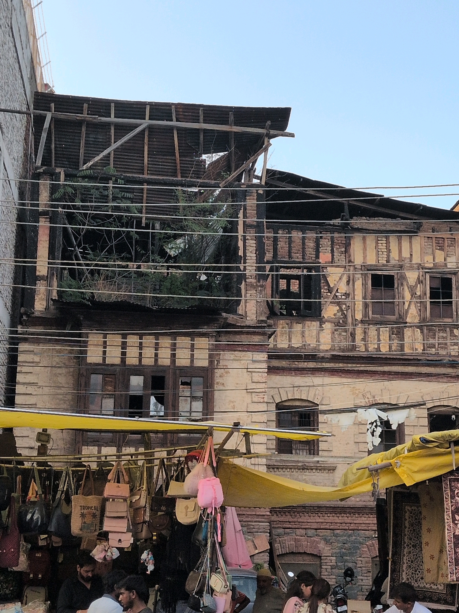 The image depicts a street scene in Srinagar, India, with a focus on a dilapidated building and a market stall. The building, on the right, appears to be old and in a state of disrepair, with exposed wooden beams and a damaged roof covered with corrugated metal. The lower floors show traditional architectural elements with multiple windows. In front of the building, a market stall is set up, displaying a variety of handbags and what appear to be textiles. Several people are visible near the stall, some interacting with each other and browsing the goods. The overall atmosphere suggests a bustling urban environment with elements of historical architecture and commercial activity.