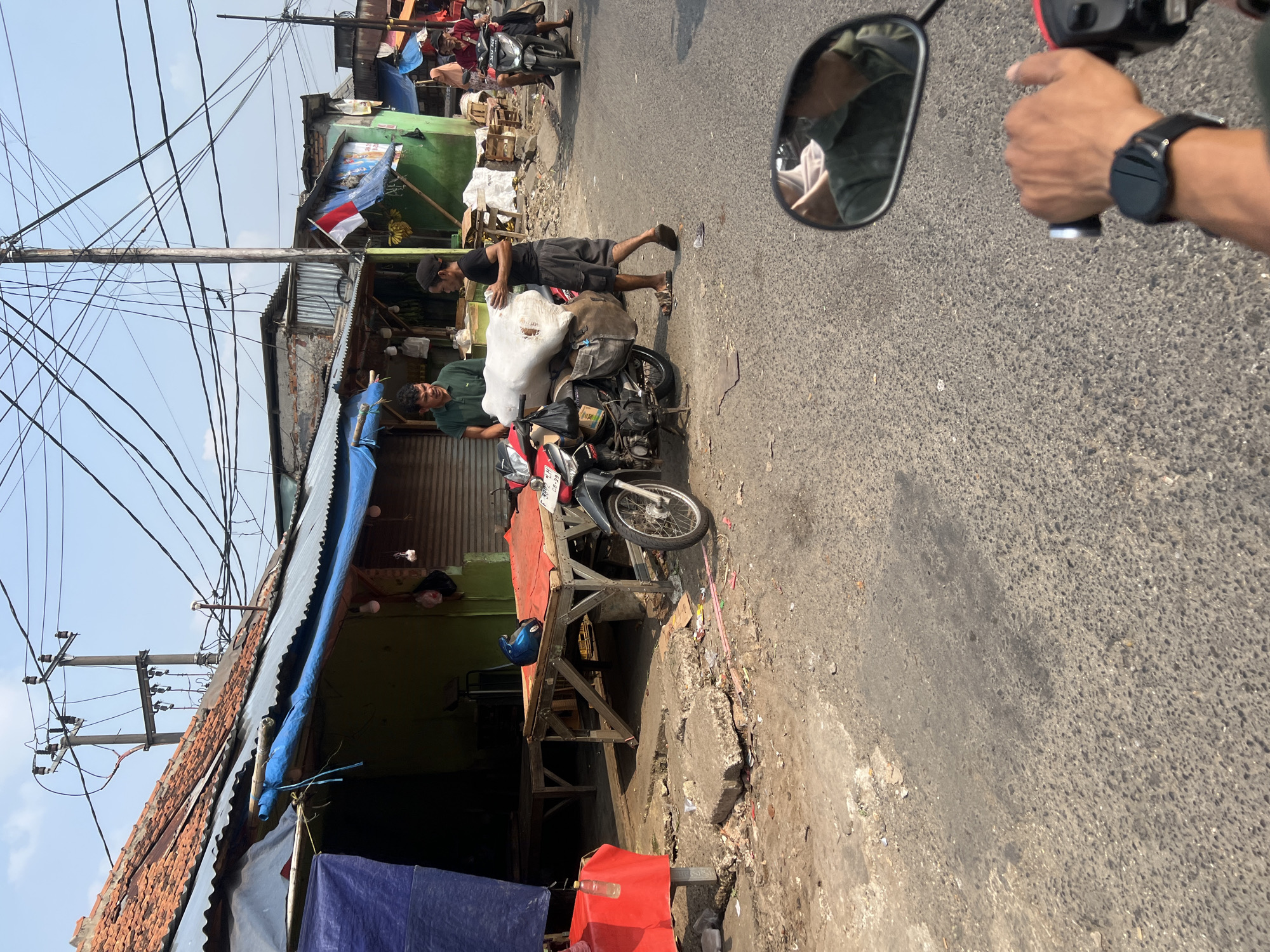 In Citeureup, Indonesia, two men are interacting next to a motorcycle loaded with a large, white, wrapped bundle. The man on the left, wearing a green polo shirt, stands near the front of the motorcycle, looking towards the other man. The man on the right, wearing a black t-shirt and shorts, is walking and carrying part of the bundle. The motorcycle, a red and black model, is parked on an asphalt road in front of a row of small market stalls. The stalls have corrugated metal roofs covered with blue tarps, and one stall displays a small Indonesian flag. The environment appears somewhat unkempt, with debris visible on the ground and numerous electrical wires overhead. In the background, another motorcycle with two people is visible, further down the road. A reflection in the mirror of a third motorcycle shows a person's arm wearing a watch, indicating the viewpoint of the photograph is from someone riding a motorcycle.