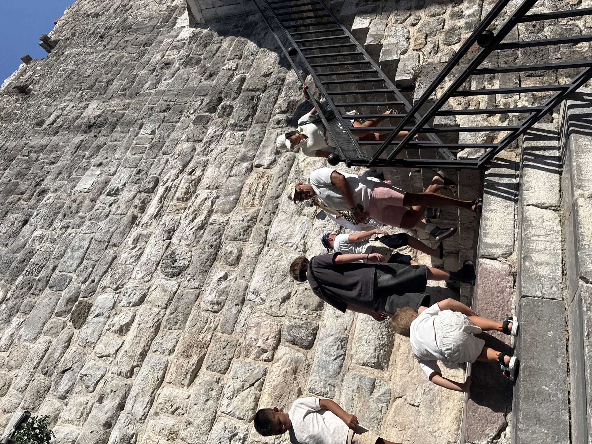 In Bodrum, Turkey, a group of people ascend a stone staircase beside a rough-hewn stone wall. A young boy in a white t-shirt and shorts crawls up a step on the left. Behind him, a young person in a dark t-shirt and shorts walks up. Further up, a man in a light-colored shirt and pink shorts, holding a saxophone, walks with a woman in a white polo shirt and skirt. Another person in white is visible further up the stairs. The scene is bathed in sunlight, casting shadows across the stone steps and wall.