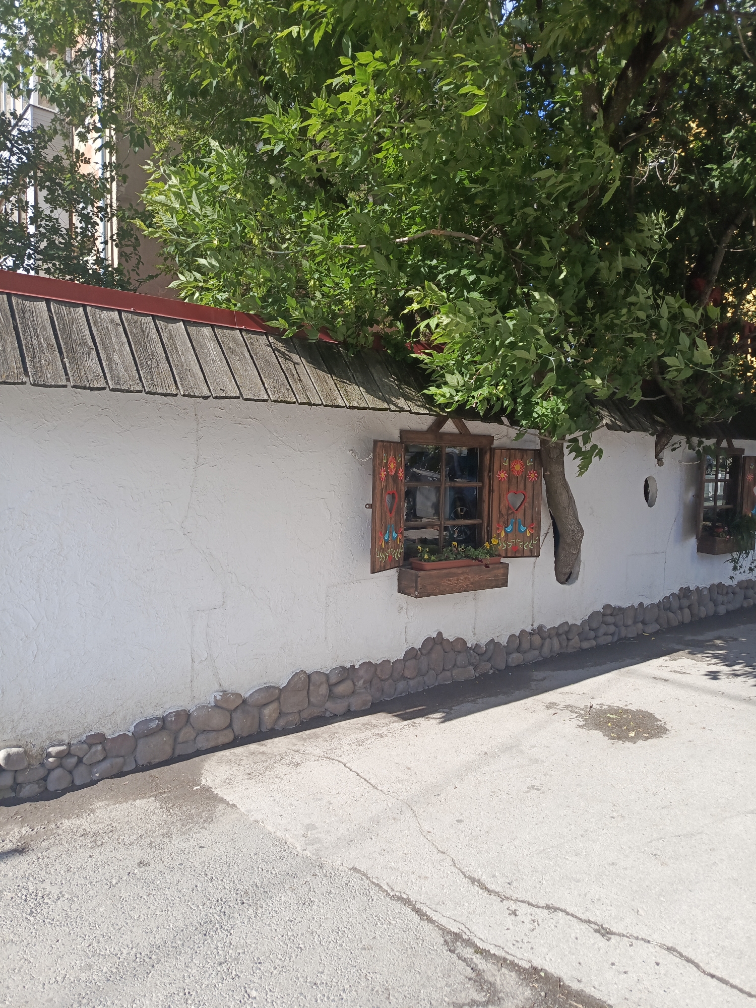 This is a medium shot, outdoor, eye-level photograph of a white plastered wall with a brown wooden, shingled roof overhang. The wall has a decorative border of smooth, rounded river stones at its base. Two windows with carved wooden shutters are visible. The shutters are painted with colorful floral designs and have heart-shaped cutouts. Flower boxes with small plants are beneath each window. A tree with lush green foliage grows through the wall, with its trunk emerging from the wall itself and its branches extending over the roof. The pavement in front of the wall is asphalt, with some cracks and a small puddle. In the background, parts of a light-colored building with windows can be seen through the tree branches. The overall scene suggests a quaint, possibly rural or traditional architectural style. This scene is located in Perm, Russia, on the street of M. Gorky.