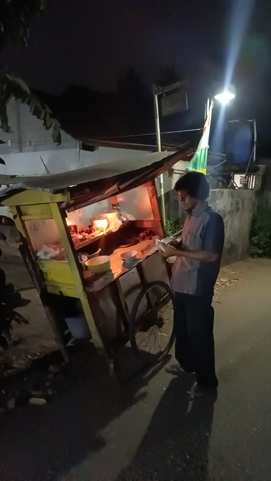 A man stands next to a food cart at night on a street in Kebumen, Indonesia. The food cart is yellow and appears to be made of wood and metal, with a covered top and a single wheel. The cart displays food items heated by red light. The man, wearing a blue patterned shirt and dark pants, is looking down at something in his hands. The street is paved and dark, illuminated by a bright street lamp overhead. Trees and a building are visible in the background. A flag with green and white stripes is attached to a pole near the street lamp. The scene suggests a typical street food vendor operating in the evening.