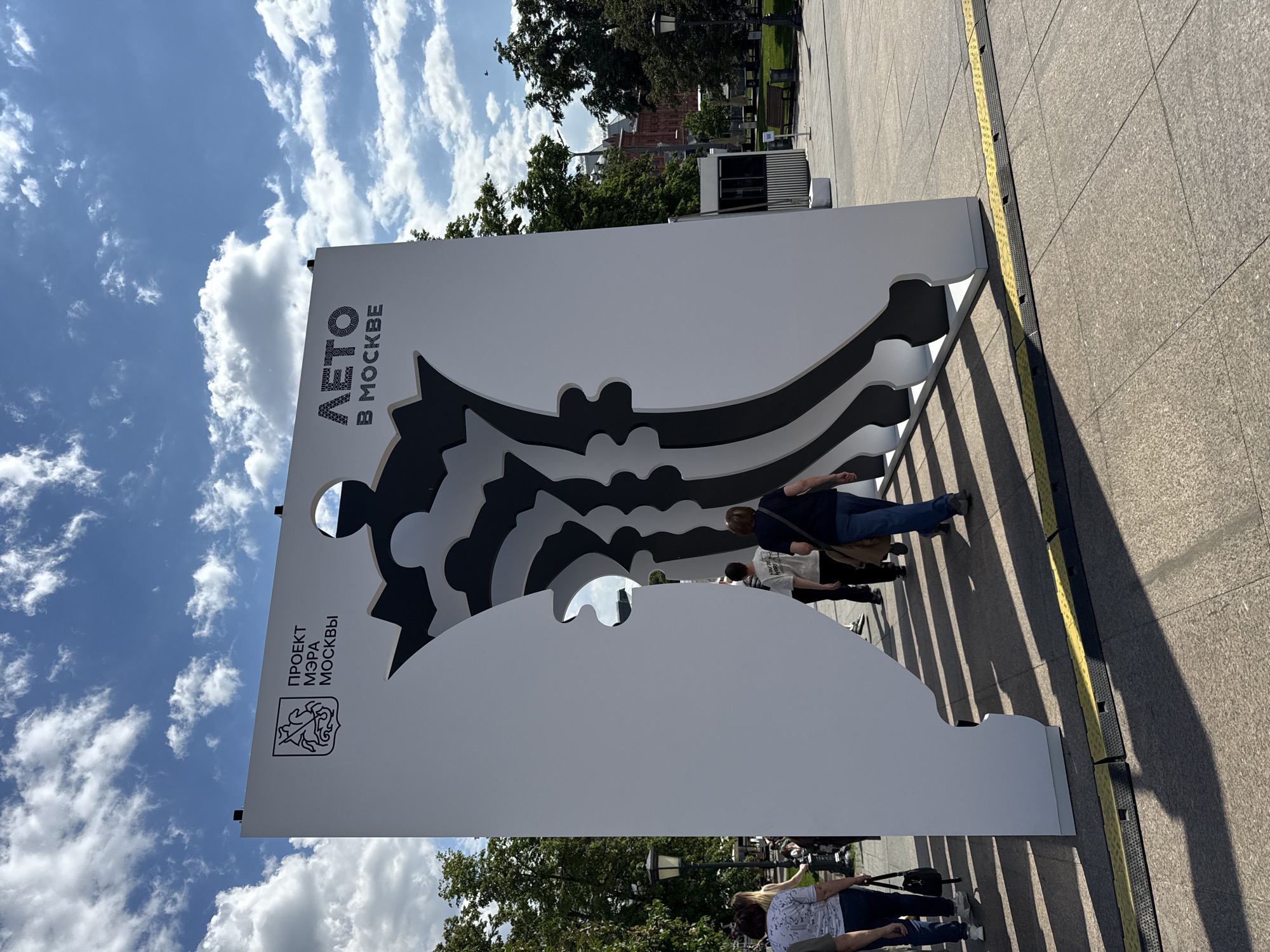A sunny outdoor scene in Moscow, Russia, features a large, white freestanding display or art installation. The structure is composed of multiple layers with dark grey and black cutout patterns that create a dynamic, wavy, and abstract design, possibly representing a cityscape or a natural landscape. Several people are visible in the background, casually interacting with or standing near the display. One person leans against the structure, looking through one of its cutouts, while others are seen walking or standing further back.
The prominent text on the display reads "ЛЕТО В МОСКВЕ" (LETO V MOSKVE), which translates to "Summer in Moscow," and "ПРОЕКТ МЭРА МОСКВЫ" (PROEKT MERA MOSKVY), meaning "Project of the Mayor of Moscow." An ornate crest or logo is positioned next to the latter phrase.
The weather is bright and clear, with a blue sky dotted with scattered white clouds. The sun is shining directly, casting long, distinct shadows from the display onto the paved ground, suggesting it is either morning or late afternoon. Trees and green foliage are visible in the background, along with hints of urban architecture, including a red brick building. The ground is a light-colored, tiled pavement with a yellow tactile paving strip running alongside the display. The overall atmosphere appears leisurely, with people enjoying the public space and the artistic installation.