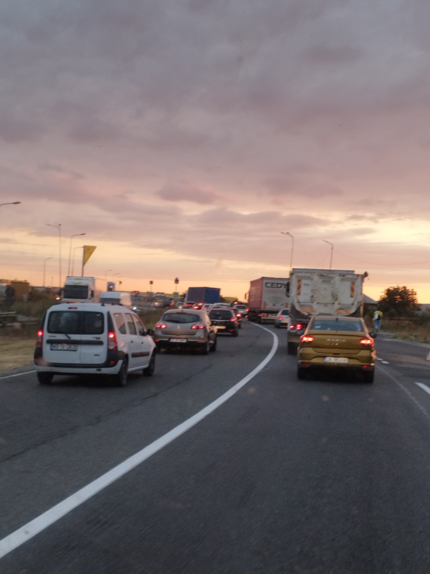 An image depicts a traffic jam on a highway during sunset in Chitila, Romania. The sky is filled with dramatic clouds in shades of pink, orange, and gray. Several vehicles are visible, including cars, vans, and trucks, all stopped or moving slowly in traffic. In the foreground, a white van with Romanian license plates is prominent. To its right, a gold Dacia sedan is also in view. Several other vehicles are further up the road, creating a sense of congestion. The road surface is asphalt with clear white lane markings. Streetlights and roadside structures are also visible in the background.