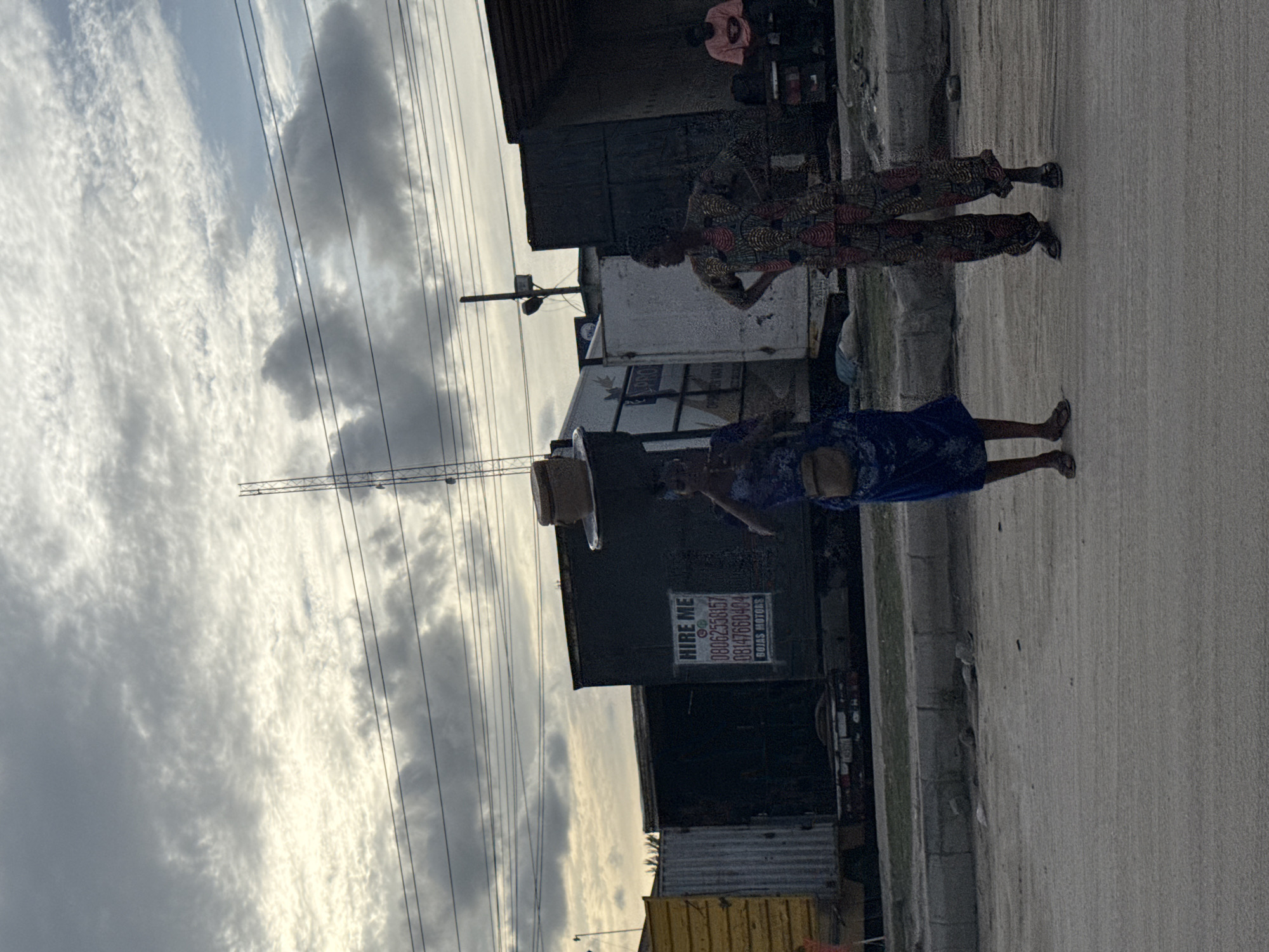 The image shows a street scene in Lagos, Nigeria. Two women are walking along a roadside. One woman is carrying a box on her head. The other is wearing a patterned top and pants. A parked vehicle with a "Hire Me" sign is visible. Overhead power lines cross the sky. The sky is cloudy. The ground is paved.