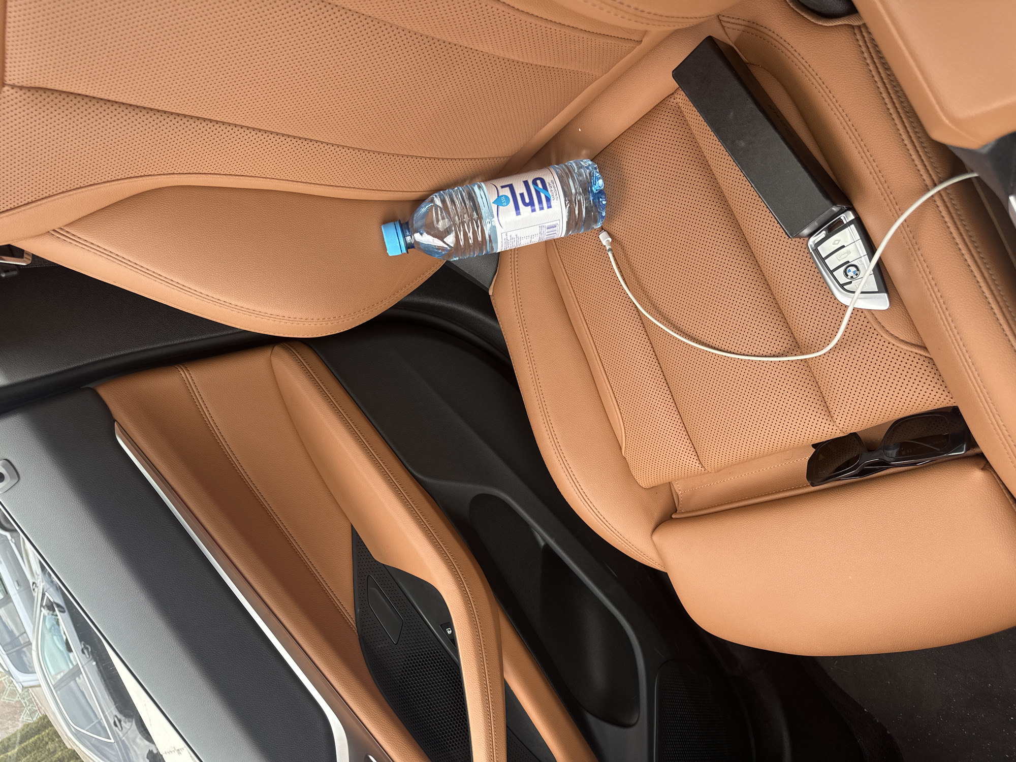 The image shows the interior of a car with tan leather seats. On one of the seats are a partially empty bottle of Fiji water, a car key fob connected to a charging cable, and a pair of sunglasses. The car door is partially visible. There is no indication of the location being Tsakhkadzor, Armenia.