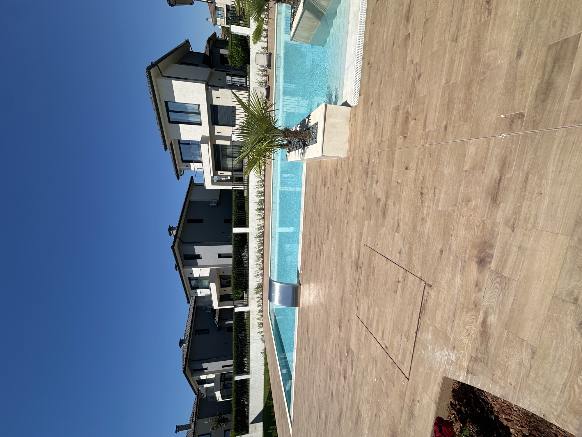 The image shows a modern residential complex in Pomorie, Bulgaria, featuring several multi-story houses with contemporary architecture, characterized by white and gray facades, flat roofs, and large windows. In the foreground, a swimming pool with bright blue water is visible, complete with a sleek stainless steel waterfall feature. The pool area is surrounded by light-colored wooden deck tiles. Two small palm trees are planted in a planter box next to the pool, with dark stones as ground cover. The sky is a clear, vibrant blue. In the distance, a black metal fence and neatly trimmed green hedges line the property. The overall scene depicts a luxurious and well-maintained outdoor living space on a sunny day.