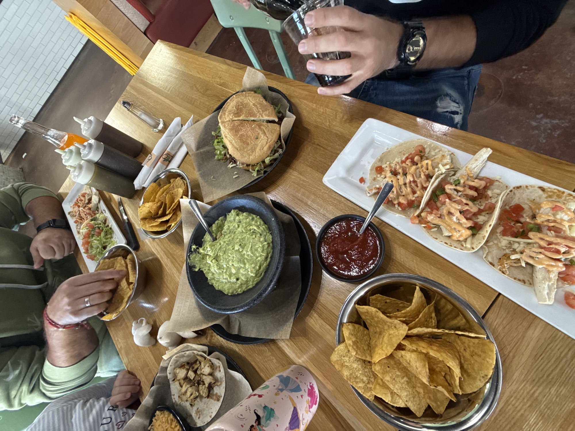 The image captures a top-down view of a wooden table laden with various Mexican-style dishes. Three individuals are partially visible around the table.
On the table, there is a platter of tacos, a sandwich cut in half, bowls of guacamole and salsa, and multiple bowls of tortilla chips. Condiments are also present. One person is pouring a dark liquid, possibly soda or beer, into a glass. Another person is eating tortilla chips with their hand.
The image does not provide enough information to confirm the location in the city of Algonquin, United States of America.