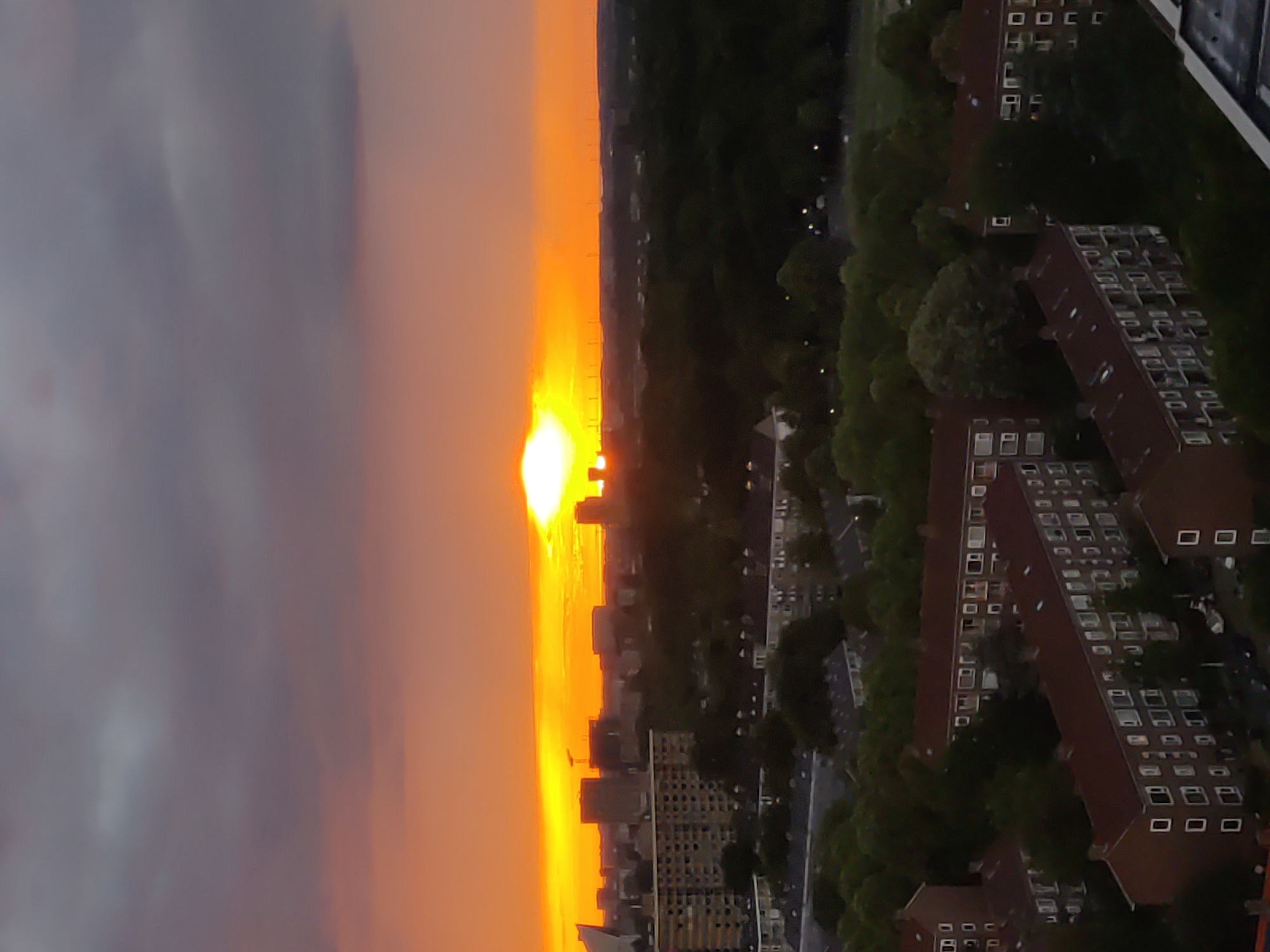 This is an aerial, high-angle, vertical shot of a city during sunset. The sky is filled with dramatic clouds in shades of purple, grey, and orange, with a bright, intensely yellow sun peeking through, casting a warm glow across the horizon. In the foreground, numerous apartment buildings with red-tiled roofs are visible. These buildings are densely packed and appear to be a residential area. The architecture suggests a European setting. The buildings are mostly rectangular, with rows of windows. Green trees are interspersed throughout the buildings, with some taller trees forming a denser canopy in certain areas. In the background, a cityscape with taller buildings and structures is silhouetted against the colorful sky, suggesting a more urban center further away. The overall impression is a twilight scene over a developed urban area, likely taken from a significant height. The location is identified as Rijswijk, Netherlands.