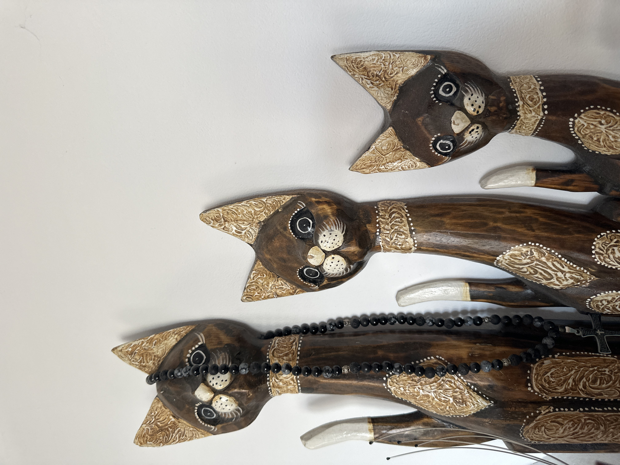 The image shows three wooden cat figurines arranged horizontally on a white surface. The figurines are brown with light-colored detailing on their heads, ears, and necks. The bottom figurine has a black beaded necklace draped across its neck, with a silver cross hanging from it. The figurines appear to be indoors, against a plain white background.