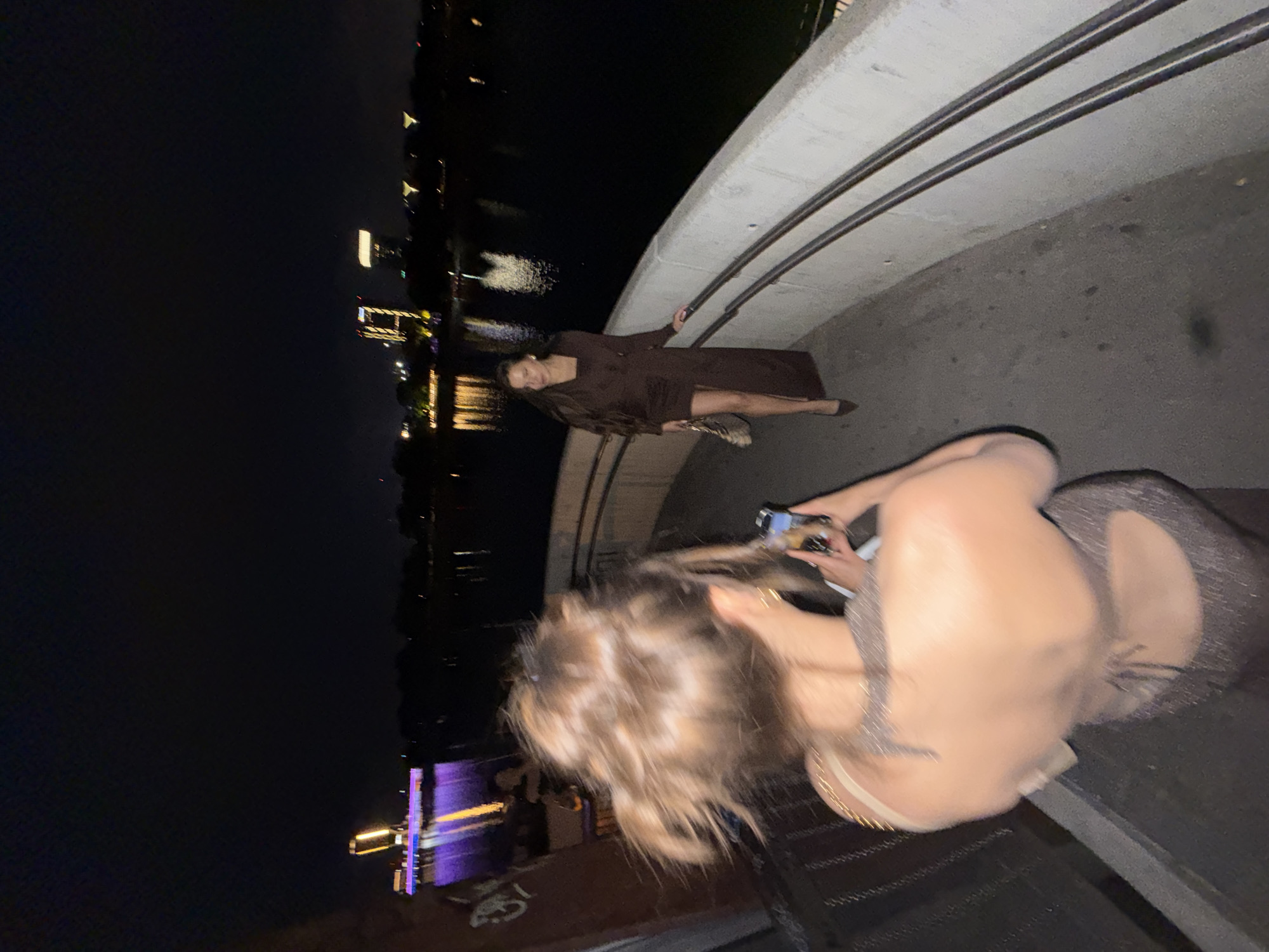 A dimly lit, slightly unfocused nighttime shot depicts two women on a walkway in Belgrade, Serbia. One woman, dressed in a brown dress, lies on the walkway. The other woman stands, holding a phone, seemingly taking a picture of the reclining woman. Buildings and their reflections are visible in the background. The scene occurs at night.