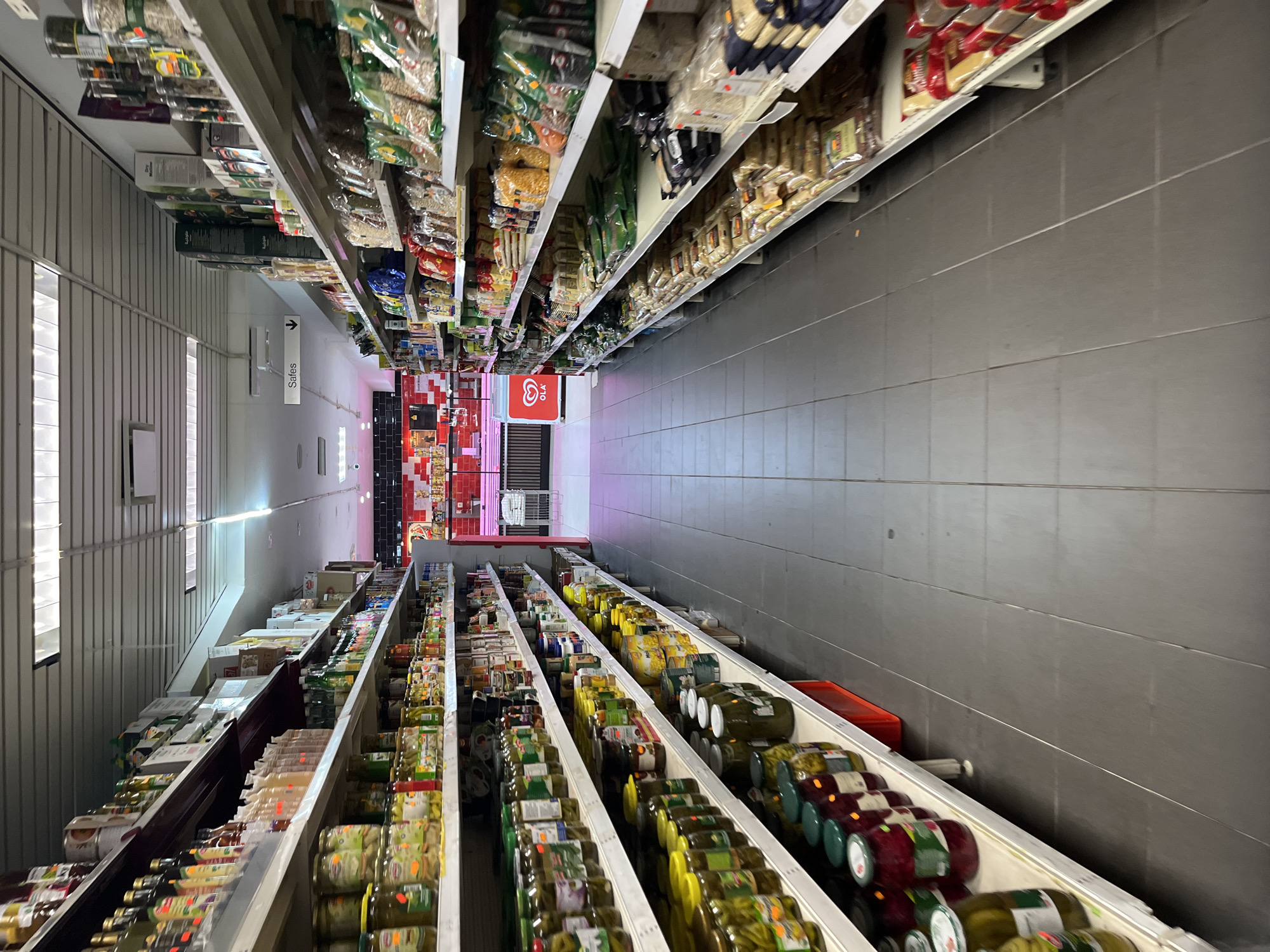 The image shows the interior of a grocery store aisle in Ostend, Belgium. The aisle is lined with shelves stocked with various food products, primarily jars of pickled vegetables and dried goods in bags. The shelves are illuminated by overhead fluorescent lighting. On the left side, jars of pickles, beets, and other preserved items are prominently displayed. On the right side, shelves are filled with packaged grains, pulses, and snacks. In the background, a red and white counter area with meat displays and a refrigerator unit with the "Ola" logo are visible. A white chair is positioned near the counter. The floor is tiled with dark grey tiles. A sign labeled "Safes" with a downward arrow is visible on the left wall. The scene depicts a typical retail environment focused on food sales.