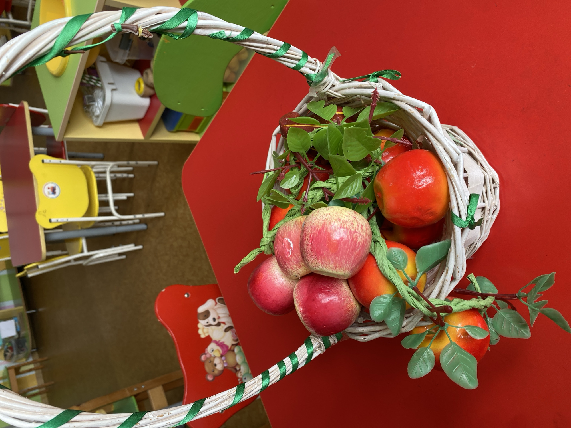 A white wicker basket filled with artificial apples and greenery sits on a red table. The basket is decorated with green ribbon. In the background, there are child-sized tables and chairs. The scene appears to be in a classroom or daycare setting. The specific location in Sochi, Russia cannot be determined from the image.