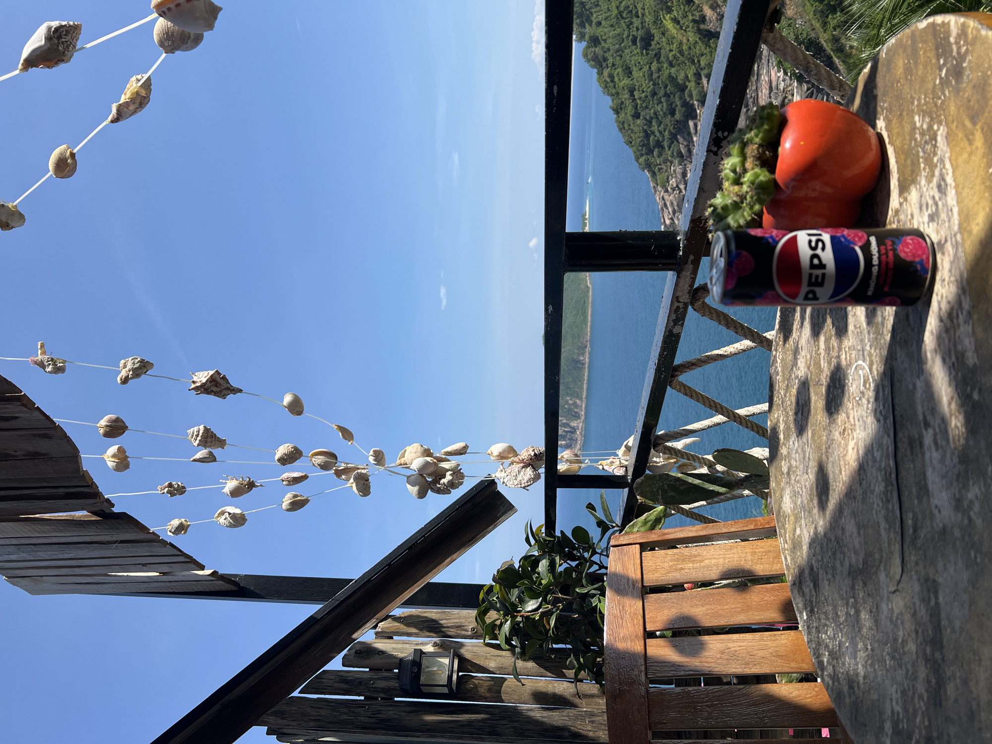From an elevated viewpoint in Hòa Xuân, Vietnam, the image captures a tranquil outdoor scene. A table, laden with a vibrant red decorative object resembling a pair of tomatoes and a cactus, and a can of Pepsi, stands beside a wooden chair. A black railing separates the table from the view of the sea and a lush, green, hilly island. Strings of seashells hang from a wooden structure above, contrasting with the clear blue sky.