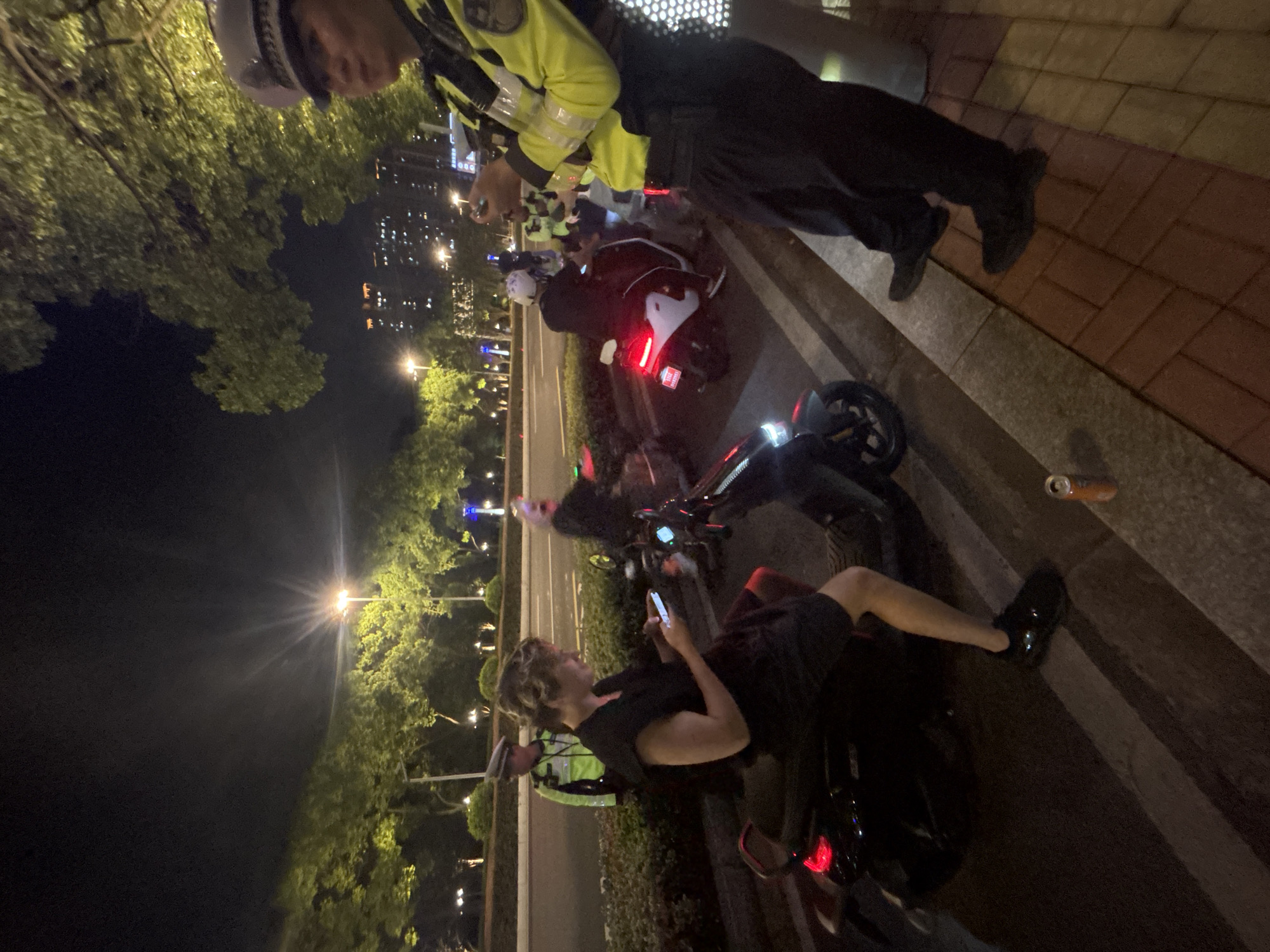 The image depicts a nighttime scene in Suzhou Industrial Park, China. A woman, dressed in a black outfit and seated on a dark-colored electric scooter, is looking at her phone. To her left, a police officer in a reflective yellow and black uniform stands, holding a phone. Behind the woman, another police officer in a similar uniform is visible standing near another electric scooter. Further in the background, more electric scooters and several trees are present, illuminated by streetlights. The environment suggests an urban setting with buildings visible in the distance, indicating the presence of the Suzhou Industrial Park. The overall mood appears to be one of casual observation or interaction between the individuals present.
