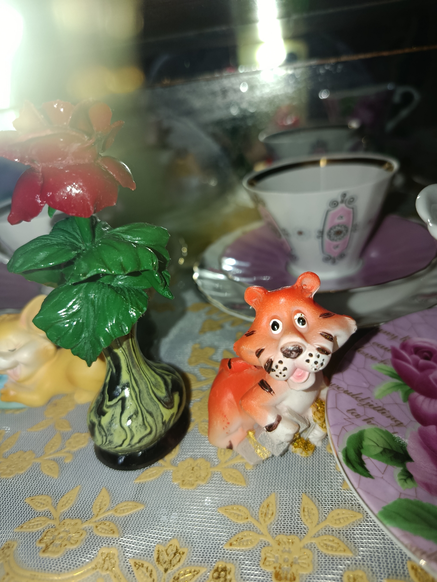This is a close-up, eye-level, indoor photograph of decorative trinkets. In the foreground, a small, orange and white tiger figurine sits on its hind legs, facing slightly to the right, with its mouth open and a gold-colored ribbon wrapped around its neck. To the left of the tiger is a small, black and green marbled vase holding a green artificial plant with large leaves. Behind the vase, a yellow, stylized rabbit figurine is partially visible. In the background, several porcelain teacups and saucers are arranged. One teacup, with a pink floral design and gold trim, is prominent. The objects are placed on a light-colored surface covered with a patterned tablecloth featuring gold floral motifs. The lighting in the image is bright and appears to come from the upper left, creating reflections and glare on the shiny surfaces of the objects. The scene is devoid of any people or discernible geographical markers that would indicate a specific location in Tashkent, Uzbekistan.