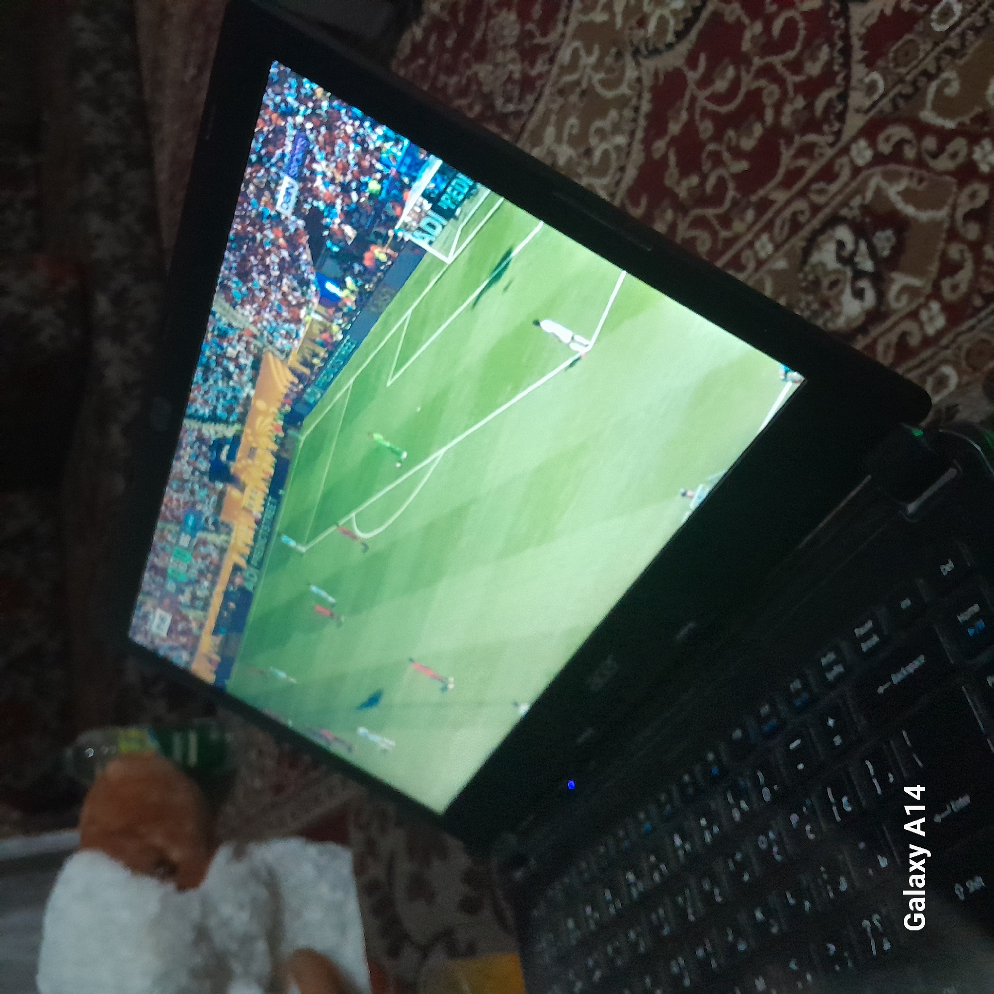 The image depicts a laptop open and displaying a football match on its screen. The laptop is positioned on a carpeted surface with a red, brown, and beige patterned design. To the left of the laptop, a stuffed toy animal, possibly a sheep, with white fluffy material and an orange face is partially visible. In the background, a dark, indistinct environment suggests an indoor setting, possibly a home, located in the city of Khan Yunis, Palestinian Territories. The laptop's keyboard is visible in the foreground, with the text "Galaxy A14" overlaid on it. A small blue light is illuminated on the laptop's body. The football match on the screen shows players in red and green kits on a green field with spectators in the stadium. The overall scene conveys a moment of leisure, with the laptop used for entertainment.
