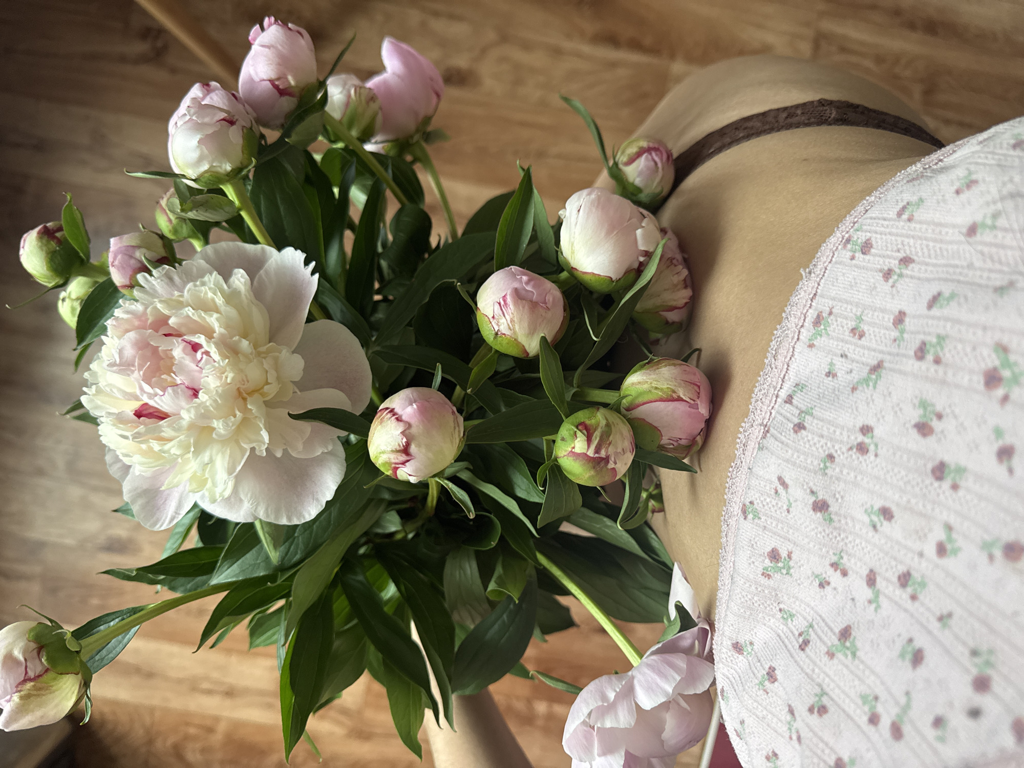 A bouquet of peonies is held by an unseen person and placed over a person's stomach and upper thigh area. The bouquet consists of one large, open white peony with pink accents and several closed pink buds. The flowers are arranged with lush green leaves. The person holding the bouquet is wearing light pink patterned underwear with a lace trim. The background consists of a wooden floor and a partially visible brown textured surface. The image is captured from a high-angle perspective.