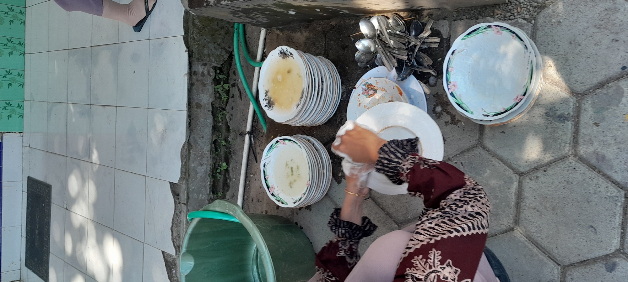 In the city of Lemahbang Dewo, Indonesia, a person is washing dishes in an outdoor setting. The ground is paved with hexagonal concrete tiles. To the left, a large green plastic basin is visible. In front of the basin, a stack of white plates with a floral rim is piled up, with some appearing to have soapy water. To the right of these plates, another taller stack of similar plates is present, also with some residual liquid. A collection of spoons and other cutlery is neatly arranged in a basket or container to the right of the plates. The person is wearing a maroon garment with a patterned design on the sleeves and is actively scrubbing a white plate with a sponge. In the background, there is a tiled wall and a patch of green vegetation. Sunlight casts shadows on the tiled ground. A green hose is also visible, extending towards the basin. A person's legs, wearing purple pants and sandals, are visible in the upper right corner of the image.