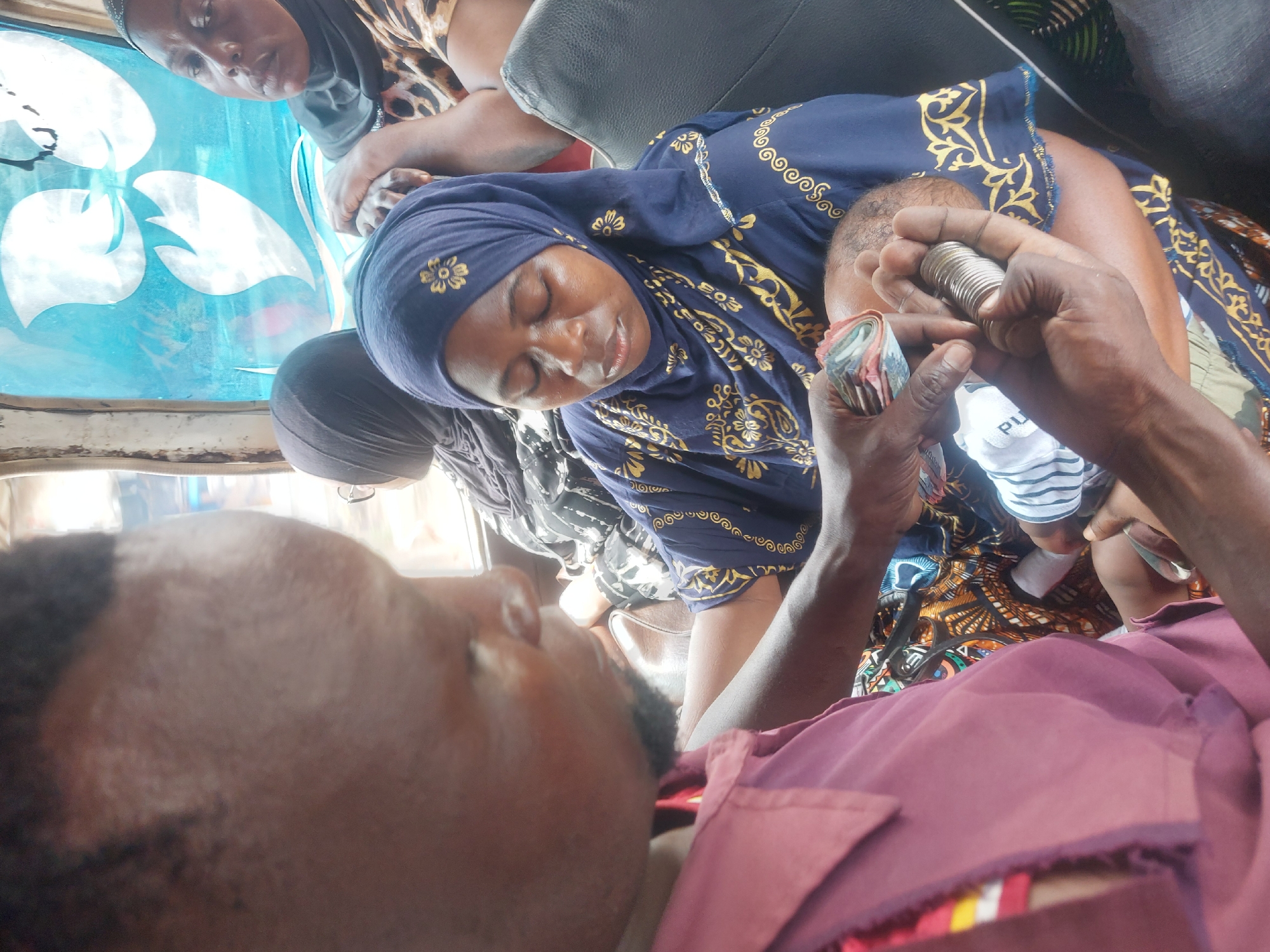 The image shows a man in a maroon shirt holding a stack of coins and rolled up bills. Next to him sits a woman in a patterned blue headscarf, who is possibly holding a baby. The scene appears to be inside a vehicle with other passengers. The exact location in Dar es-Salaam, Tanzania, cannot be determined from the image.