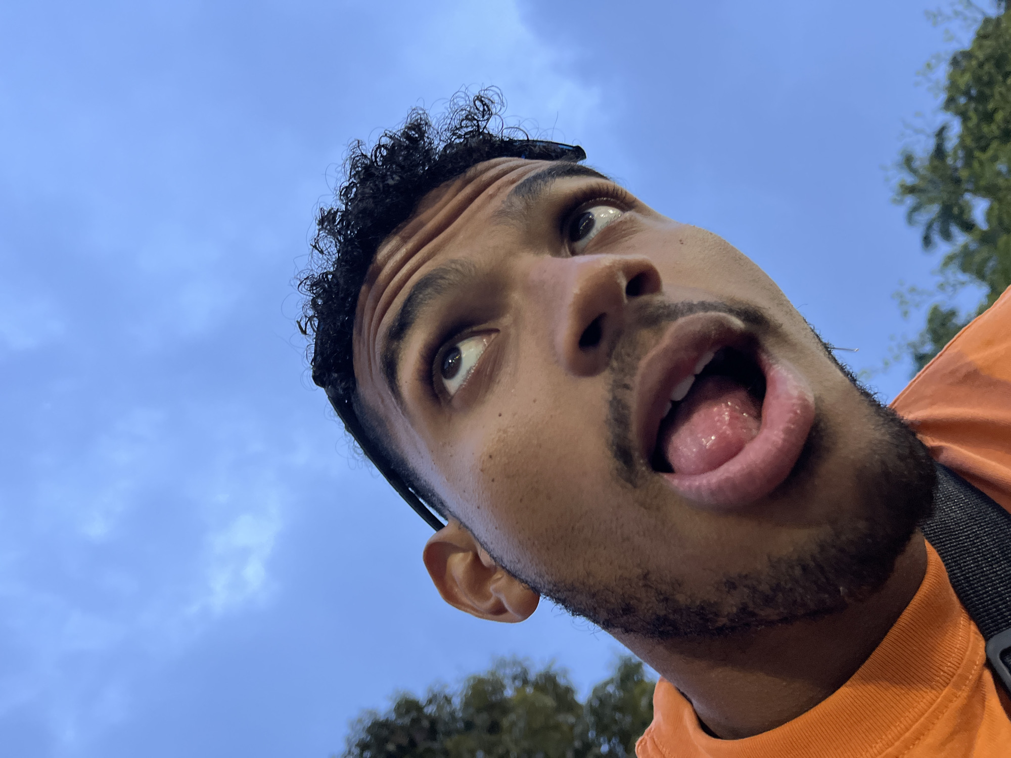 A man's face is captured from a low angle against a blue sky with scattered clouds. He is wearing an orange shirt and has curly dark hair. His mouth is open, and his tongue is sticking out. The background includes some trees and foliage, suggesting an outdoor setting. The image does not contain any information that would allow for identification of the location in the city of Mérida, Venezuela.
