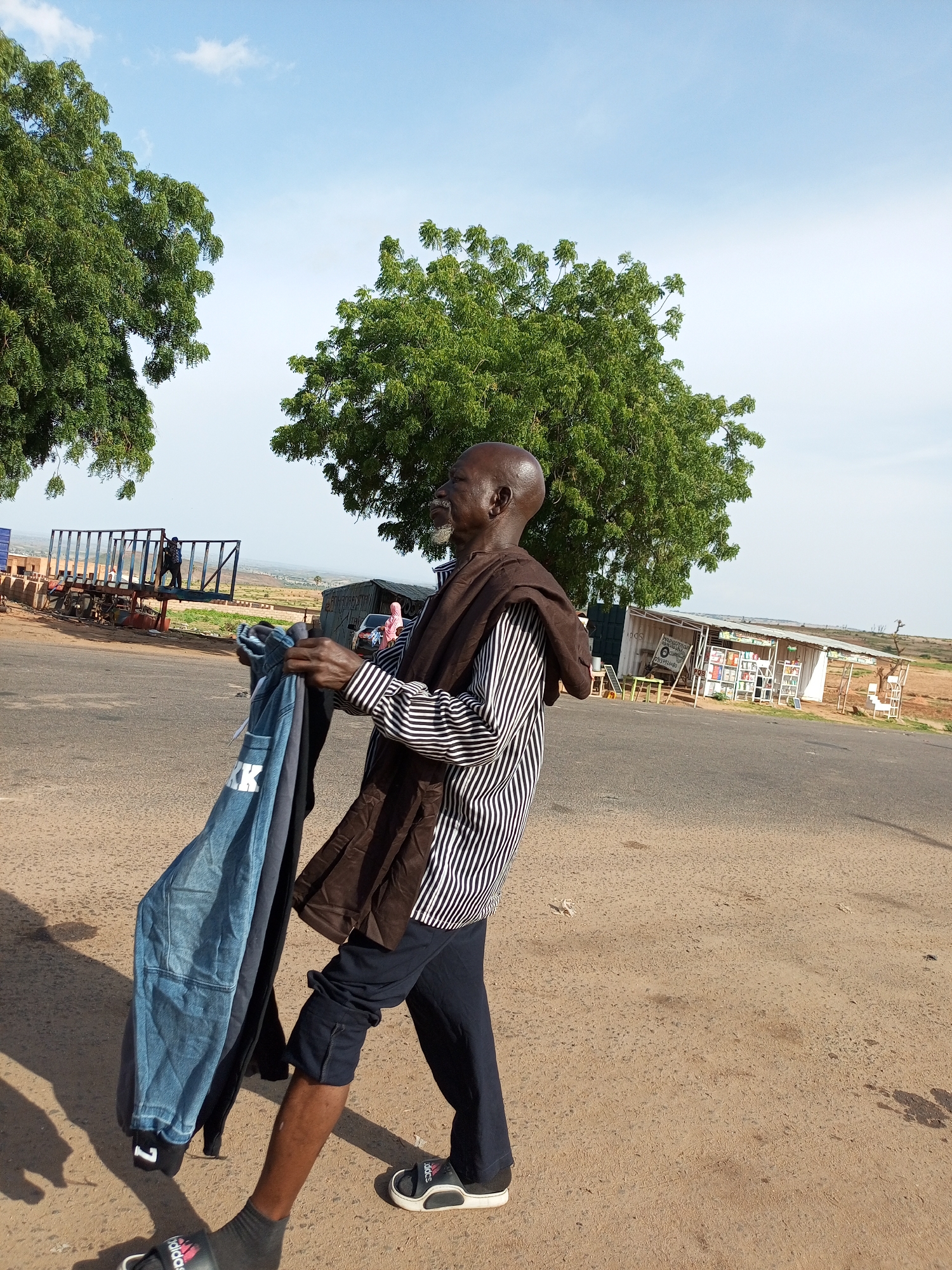 In Gadao, Nigeria, an elderly man with a bald head and a light beard walks on a dusty road. He is wearing a black and white striped long-sleeved shirt, dark trousers, dark socks, and black and white slide sandals. He is carrying a bundle of clothes in his left hand. The clothes include a blue denim garment with the letters "KK" printed on it, and another dark-colored garment. He is draped with a brown cloth over his shoulders and arms. The background shows a few trees, a large trailer, and a small roadside stall with goods displayed. The sky is partly cloudy with a light blue hue.
